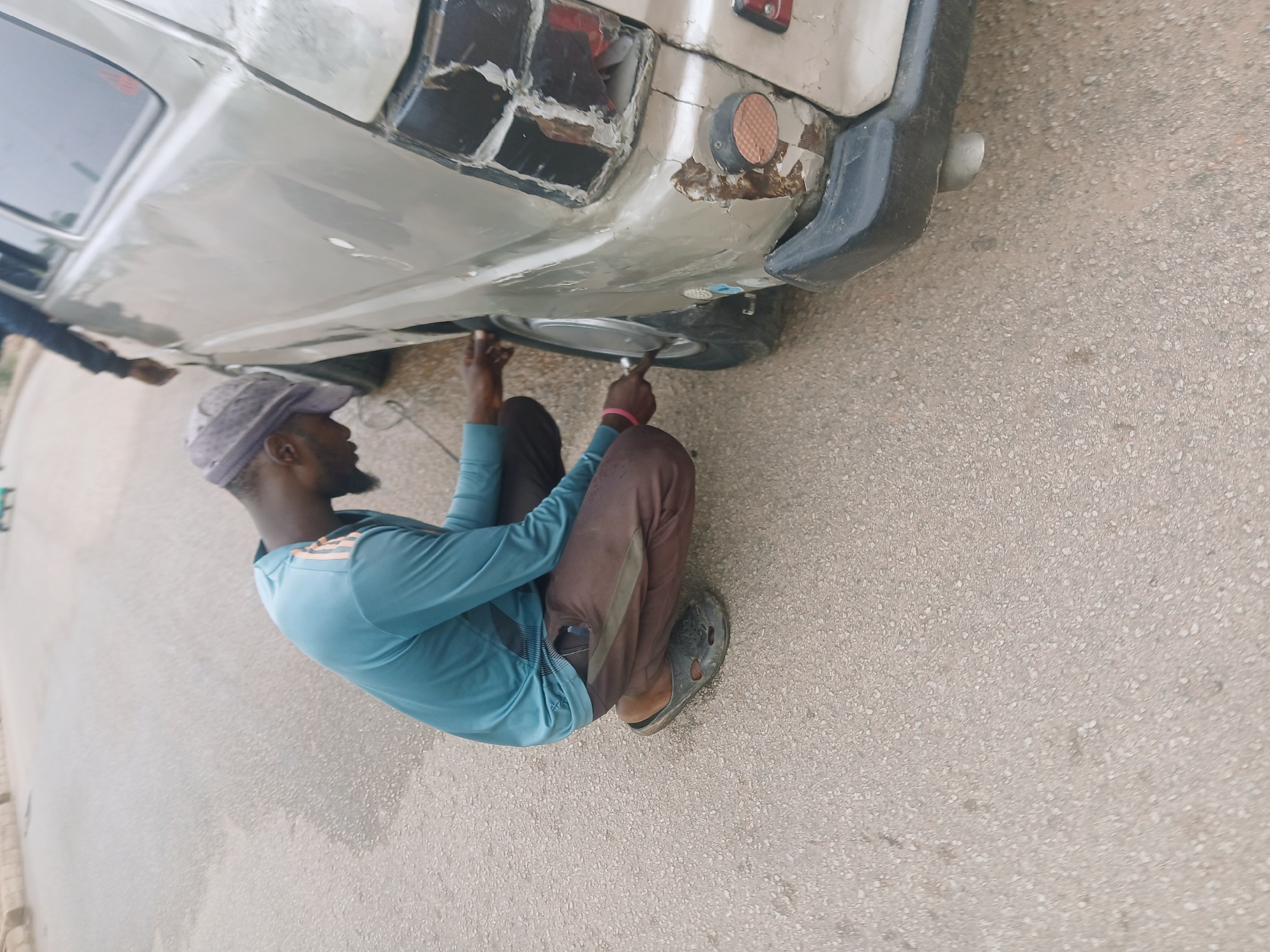 The image depicts a man in Bukuru, Nigeria, working on the rear of a light gray vehicle on a paved surface. The man is wearing a light blue long-sleeved shirt, dark pants, a hat, and slip-on sandals. He is crouched down and appears to be using a tool, possibly a wrench, on the vehicle's undercarriage near the rear tire. The vehicle has visible damage to its rear, including a broken taillight. The background shows a street.