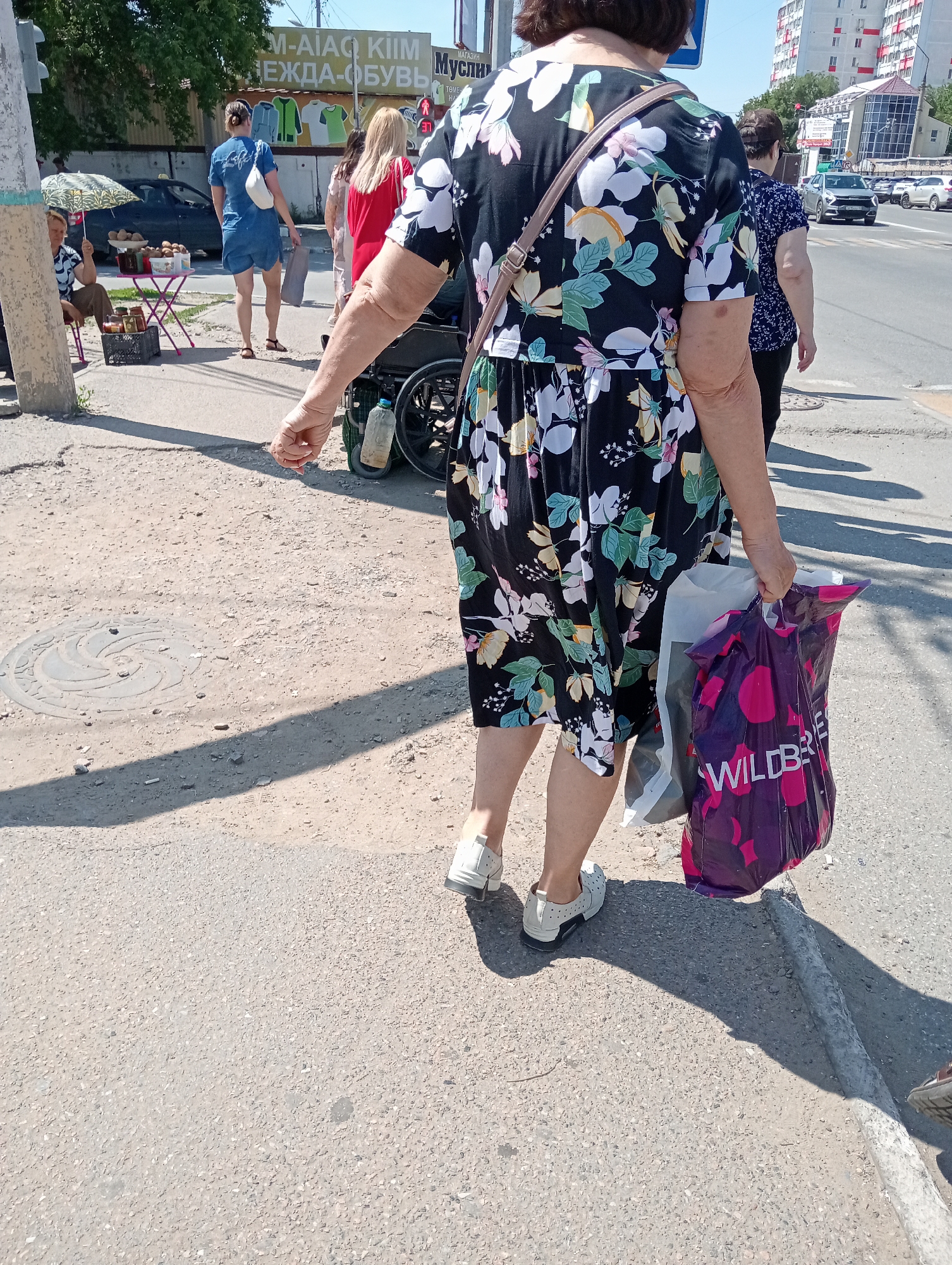 An eye-level, medium shot captures a woman from the waist down as she walks on a sunlit, dusty sidewalk in an urban environment. She is wearing a black dress with a floral pattern of white, green, and yellow flowers and leaves, paired with white lace-up shoes. Her right arm is bent, and she is carrying two shopping bags, one purple with pink polka dots and white text that reads "WILDBERRIES," and another white bag. A portion of a wheelchair is visible behind her. In the background, other individuals are walking, including a woman in a blue denim outfit with a white fanny pack and a woman in a red top. Further back, a parked car, trees, and buildings are present. The ground is paved with asphalt and dirt, with a circular metal manhole cover visible in the foreground. The scene suggests a shopping or recreational outing in an outdoor market or street.