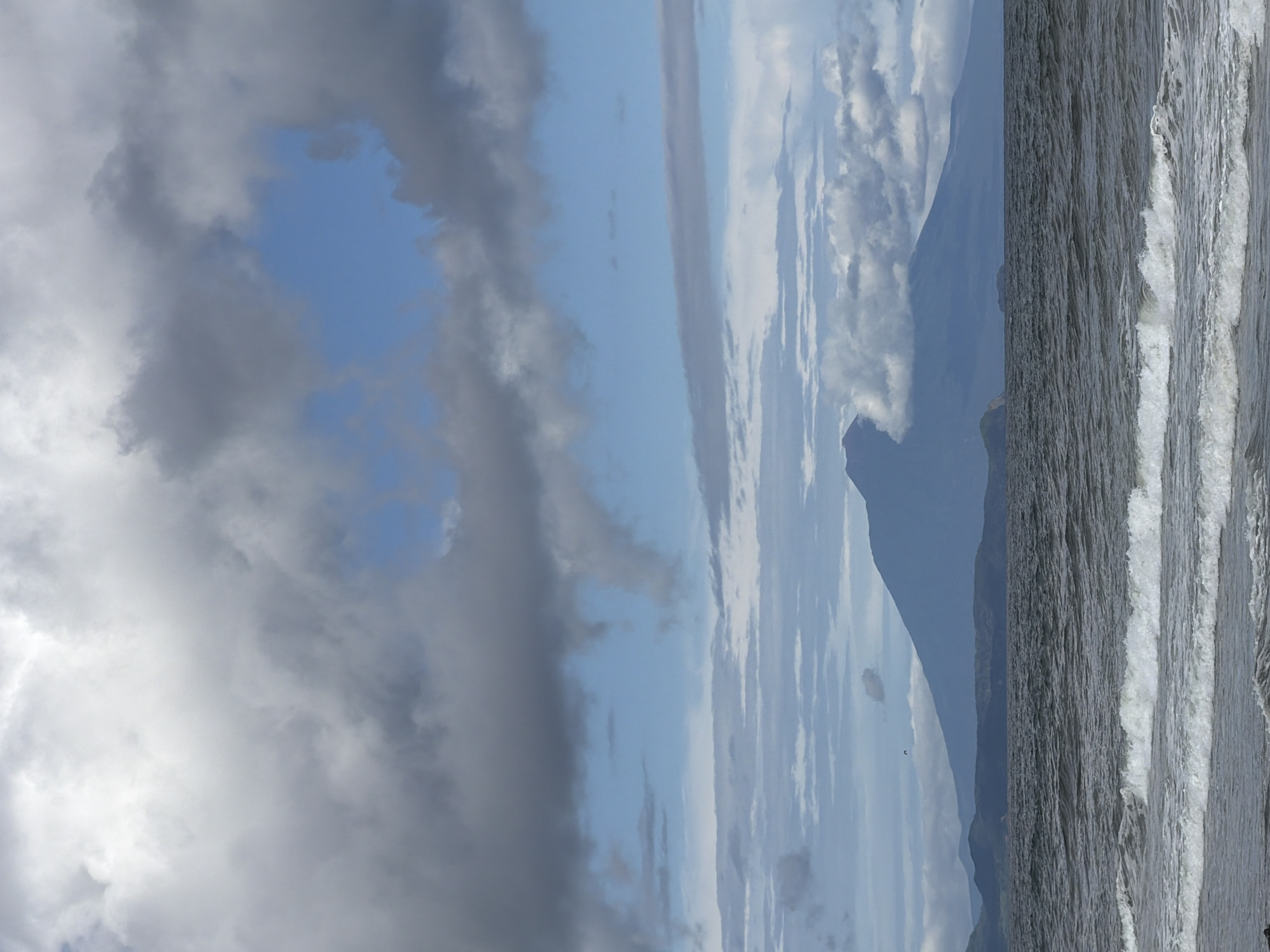 The image depicts a view of the sea and a distant mountain range under a cloudy sky. In the foreground, waves are breaking on a dark, textured surface, likely the shore. The sea is choppy with visible whitecaps. The middle ground shows a dark body of water stretching towards a hazy horizon. Behind the water, a mountain range is visible, with the peak of a large, conical mountain partially obscured by clouds. The sky is filled with a mix of dark grey and lighter grey clouds, with patches of blue sky visible in between. A single, small dark shape, possibly a bird or a kite, is discernible in the sky above the mountains. The overall atmosphere is overcast and somewhat dramatic. The location is Yuzhno-Kurilsk, Russia.