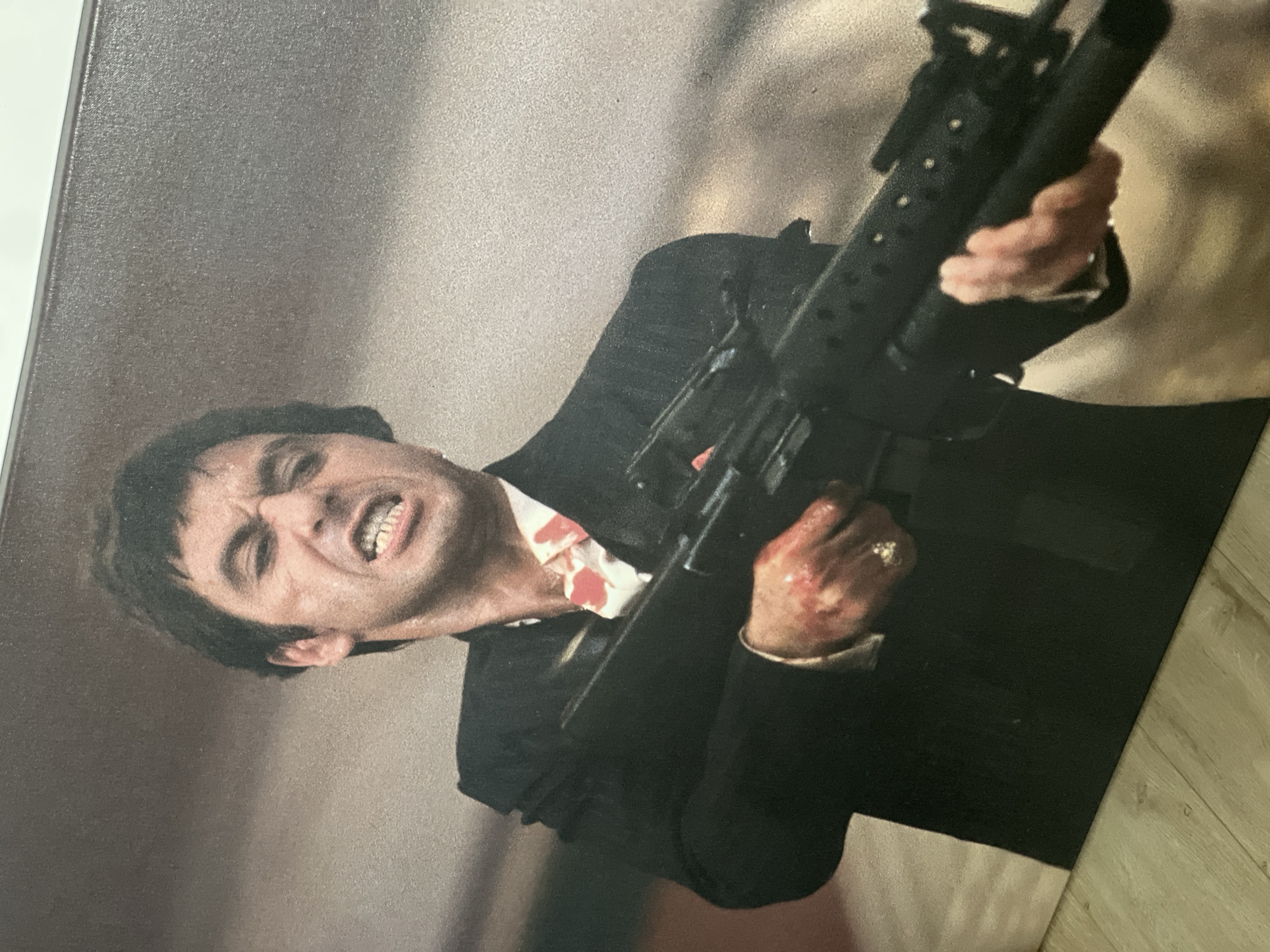 A print of Al Pacino as Tony Montana from the movie "Scarface" is shown. He is holding an assault rifle and is visibly angry. He is wearing a black suit and white shirt with bloodstains. The background is a blurred tan color. The print is laying on a light-colored wooden surface. The location is not identifiable as Vologda, Russia.