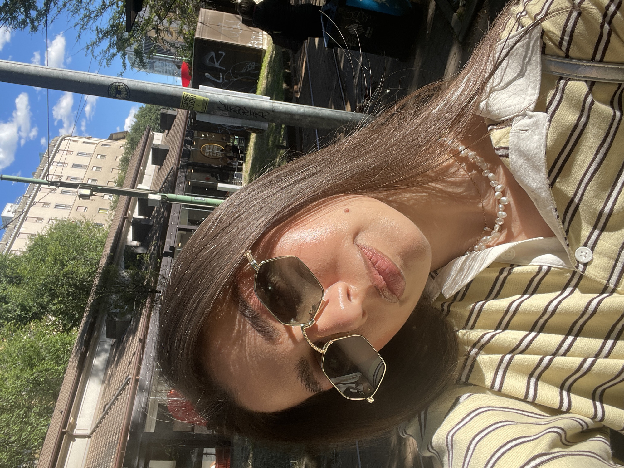 The photograph depicts a young woman with brown hair, wearing sunglasses and a pearl necklace, leaning into the camera. She is wearing a striped yellow and white collared shirt. The background shows a cityscape, including buildings and trees, suggesting an urban environment. A pole with signage is visible in the background. Based on the architectural style and urban layout, the photograph was taken in Milan, Italy.