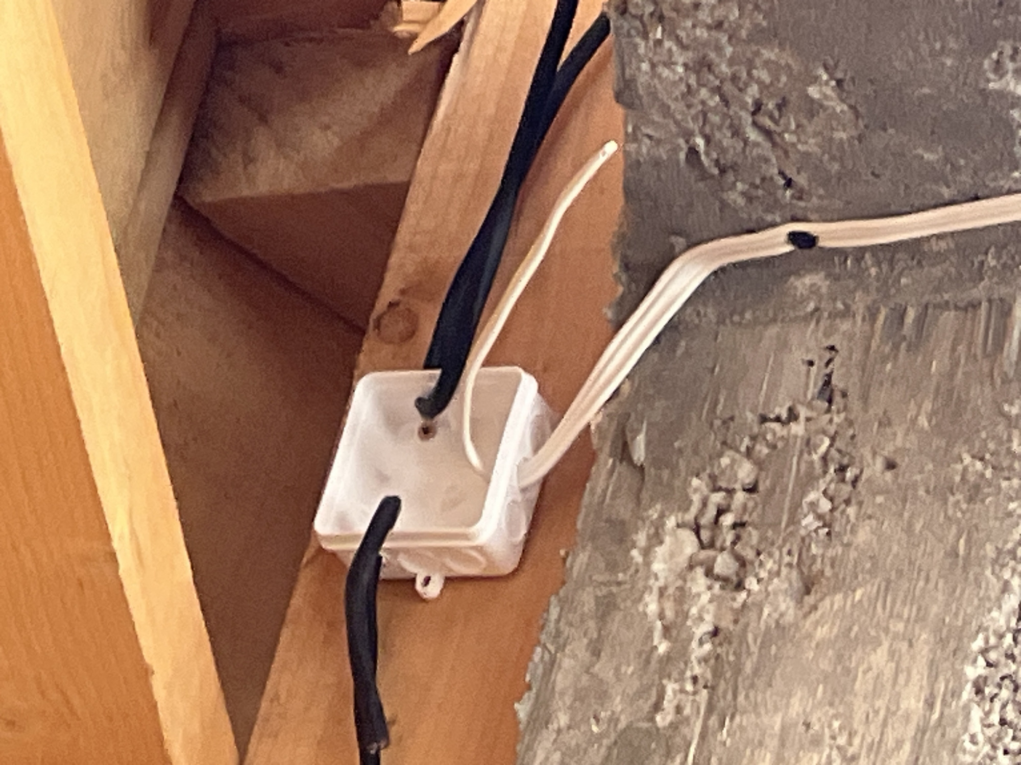 The image shows a white plastic electrical junction box mounted on a wooden beam within a structure, possibly an attic or unfinished area. Two black cables and one white cable enter and exit the box. A second white cable runs along a rough, textured wall. The scene appears to be indoors. Location of the scene is unknown.