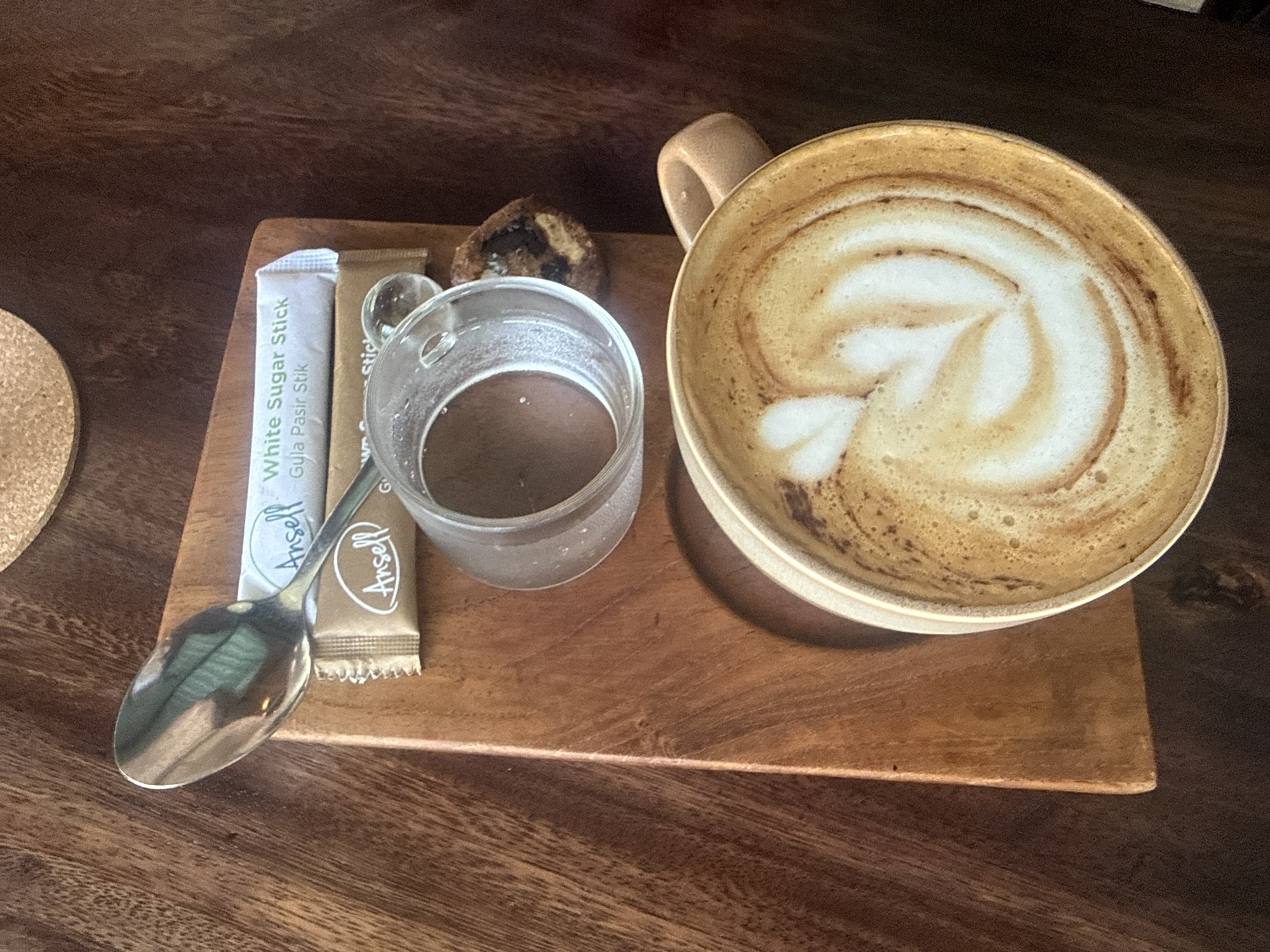 The image presents a close-up, top-down view of a coffee service set on a wooden table, likely in a cafe in the Ubud District, Indonesia. The main subject is a light-colored ceramic cup on the right, filled with a latte or cappuccino adorned with delicate white latte art, resembling a heart or leaf, against the creamy froth and coffee.
To the left of the coffee cup, a small, clear glass holds a dark brown liquid, potentially cold brew coffee or water. Adjacent to the glass are two sugar stick packets: a white one clearly labeled "White Sugar Stick Gula Pasir Stik Ansell" and a brown one also displaying "Ansell." A polished silver spoon rests partially over these packets, reflecting parts of the room. Behind the glass and sugar sticks, a small, dark brown pastry or cookie with lighter speckles is visible.
All these items are neatly arranged on a rectangular wooden serving tray, which in turn sits on a larger, darker brown wooden table. A circular cork coaster is partially visible on the far-left edge of the frame. The lighting is soft and diffused, suggesting a daytime setting. The visible Indonesian text "Gula Pasir Stik" (meaning "Sugar Stick") on the sugar packet serves as a location cue, confirming the given context of Indonesia. No people are visible, and the scene conveys a peaceful, inviting cafe ambiance.