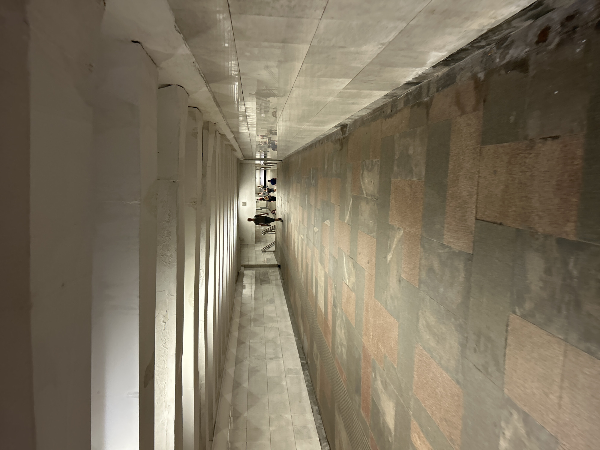 An overhead shot shows a wide, rectangular underground pedestrian tunnel. The tunnel walls are tiled in a light, off-white color, and the ceiling is constructed of white concrete beams. The floor is paved with large, rectangular tiles in a pattern of alternating reddish-brown and dark gray colors. Several people are visible in the tunnel. In the foreground, a single person walks away from the camera toward the far end of the tunnel. Further down the tunnel, a group of people are gathered, and another individual walks toward the viewer. Reflections of people and the tunnel structure are visible on the glossy wall to the right. The tunnel appears dimly lit, with light sources unseen. The environment is clean but shows signs of wear, with some water staining at the base of the right wall.