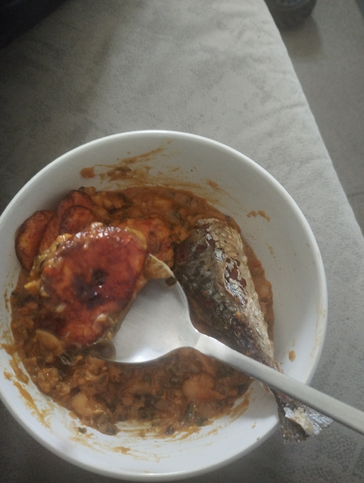 The image shows a white bowl filled with food, likely sitting indoors. The bowl contains what appears to be beans stew, fried plantains, and a whole fried fish. A metal spoon is partially submerged in the food. The location is claimed to be Dutsen Alhaji, Nigeria, but this cannot be independently verified from the image. No people are visible.