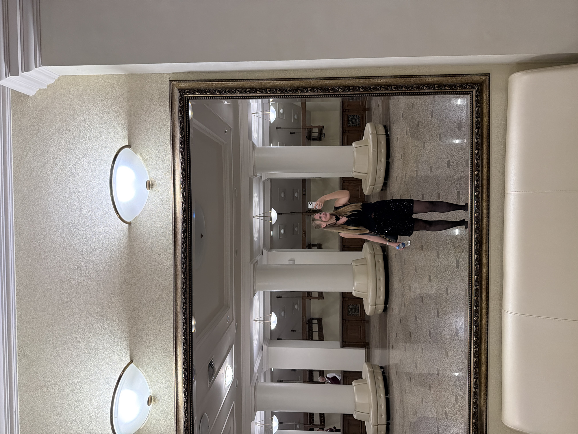 A woman stands in front of a large mirror in an ornate frame. The mirror reflects a long hallway with white columns and built-in seating. The woman is holding a cell phone and appears to be taking a selfie. She is wearing a black dress and dark tights. The location is likely a public space such as a hotel lobby or reception area. The city of Izhevsk, Russia is not confirmed through visual elements. The scene includes lamps mounted on the wall near the mirror.