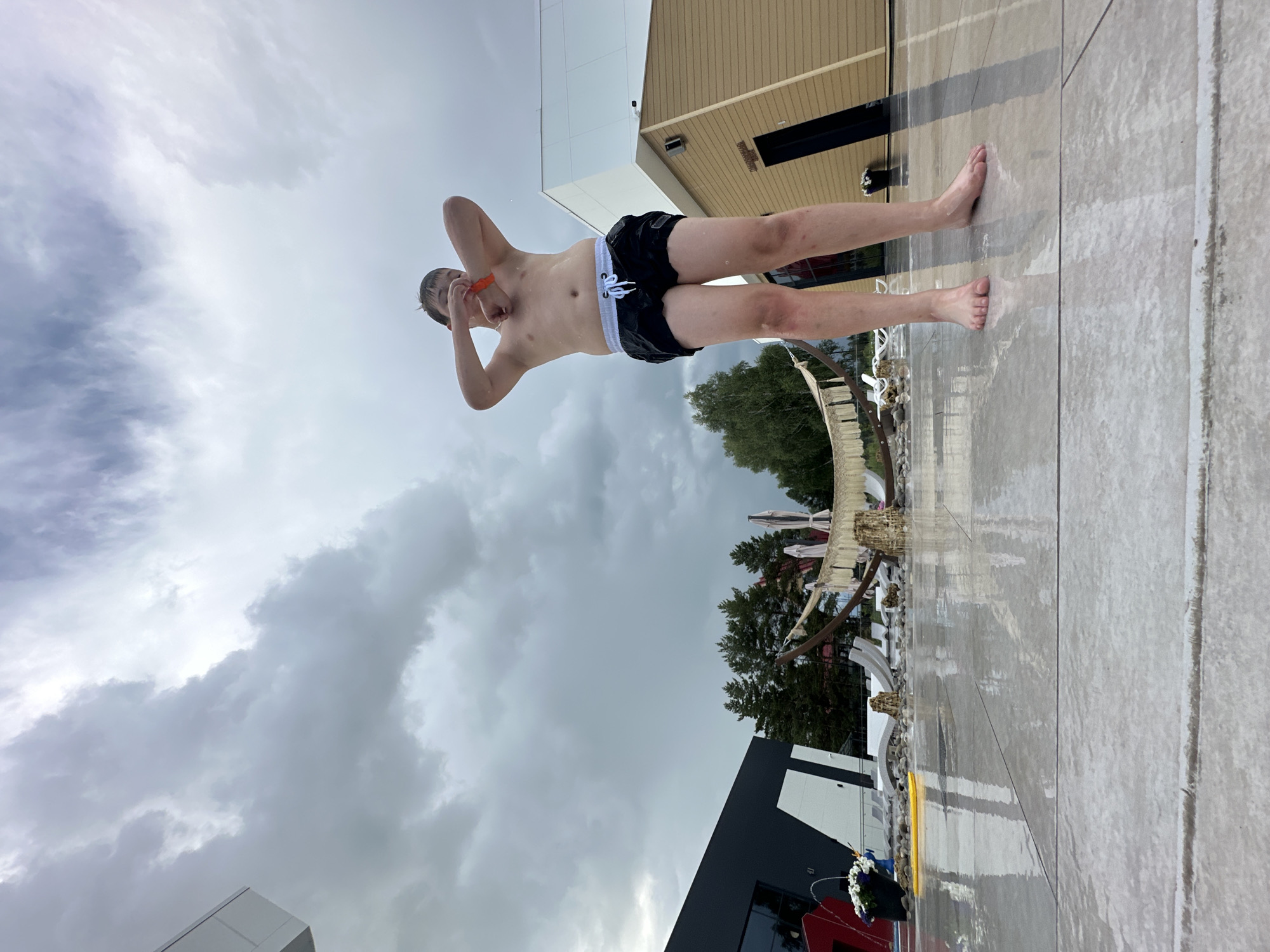 A young person in black swim trunks stands with their hands behind their head, looking upwards towards a cloudy sky. They are standing on a wet, tiled surface, possibly a poolside. Behind them, a building with beige siding and a modern black and white section is visible, along with trees. The sky is overcast and appears to be either dawn or dusk, or perhaps a very stormy day. The person's skin is wet, suggesting they have recently been in water. There is no visible text in the image.