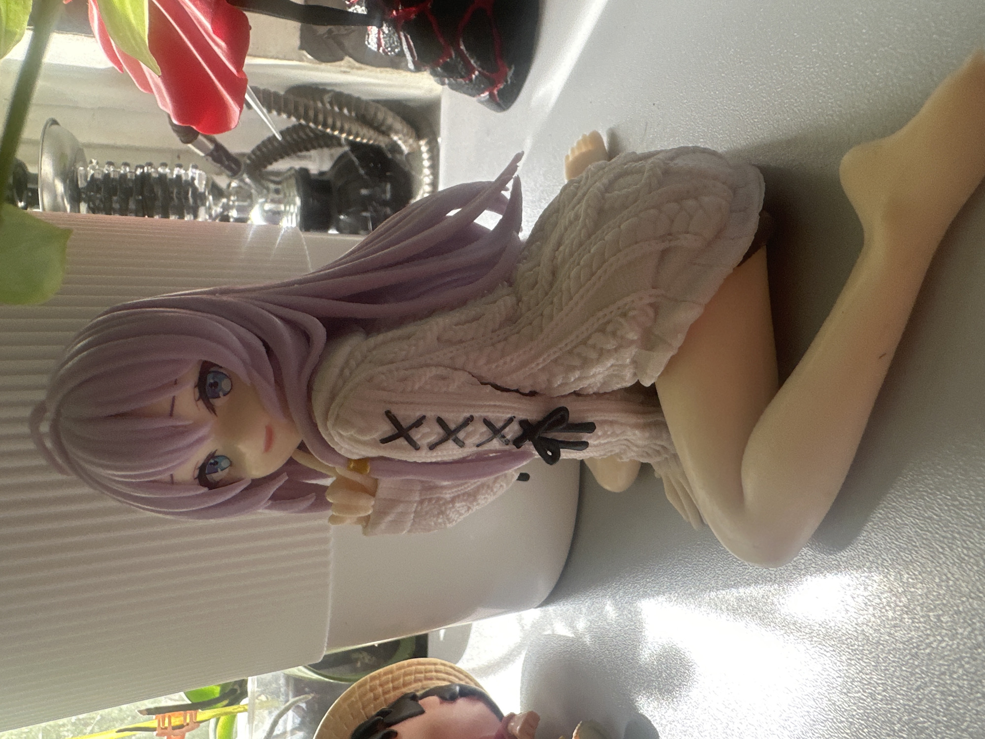 An anime-style figurine of a young woman with lavender hair and blue eyes is kneeling on a gray surface. She is wearing a chunky knit white sweater with criss-cross stitching details on the sleeve and a black bow. Her right leg is bent, with her foot visible, while her left leg is extended forward. Sunlight casts shadows and highlights on the surface and the figurine. In the background, a white ribbed pot or planter is to the left, with greenery visible behind it. To the right, a silver hookah with a black hose is partially visible, along with a red decorative object. The scene is indoors, with a window in the background. The location is stated to be Maladzyechna, Belarus.