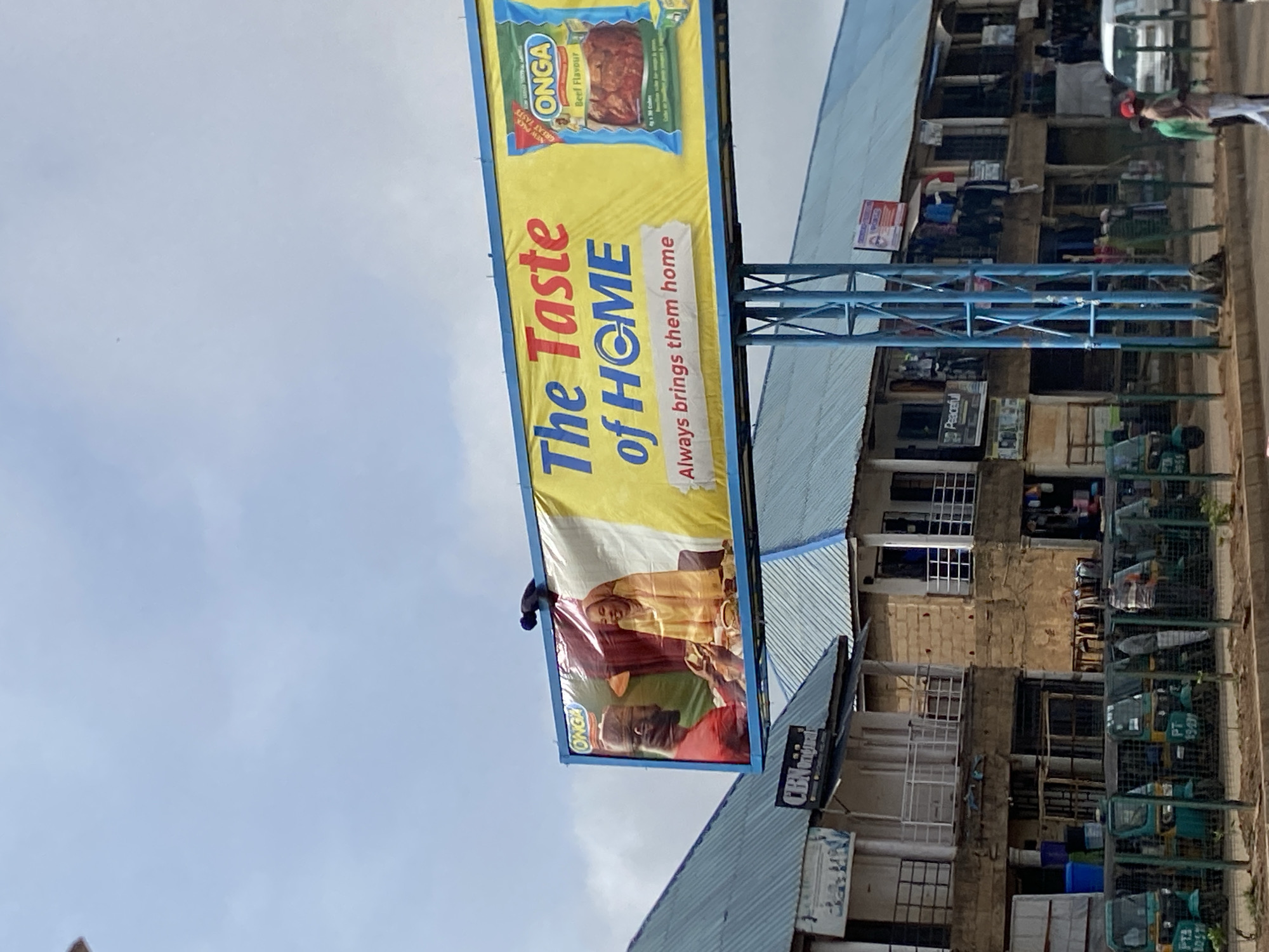 The image depicts an outdoor advertisement for ONGA Beef Flavor. The sign, which reads "The Taste of HOME. Always brings them home," features a picture of a smiling family enjoying a meal. The building in the background appears to be commercial with clothing hanging from windows. Parked along the street on the right are tricycles, used for transportation. It appears to be in a densely populated area of Bukuru, Nigeria.