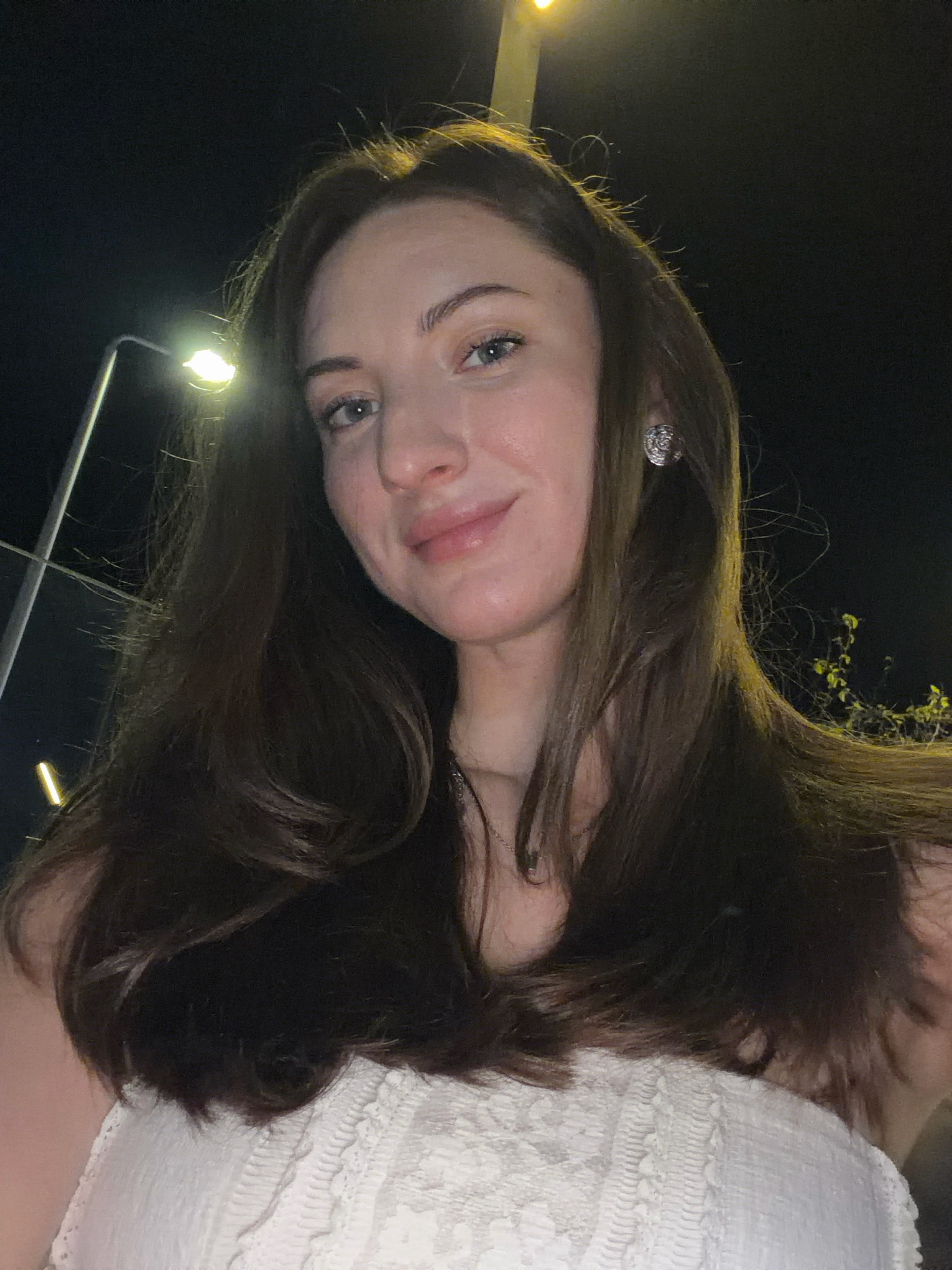 The image shows a woman with long, dark brown hair and fair skin, wearing a white top with lace detailing. She has blue eyes and is wearing a silver earring in her right ear. She is illuminated by streetlights, suggesting she is outdoors at night. The background includes streetlights and some foliage, consistent with an outdoor urban setting. The image composition suggests a selfie taken from a low angle. The scene is consistent with a location in Sharm El Sheikh, Egypt, known for its vibrant nightlife and street lighting.