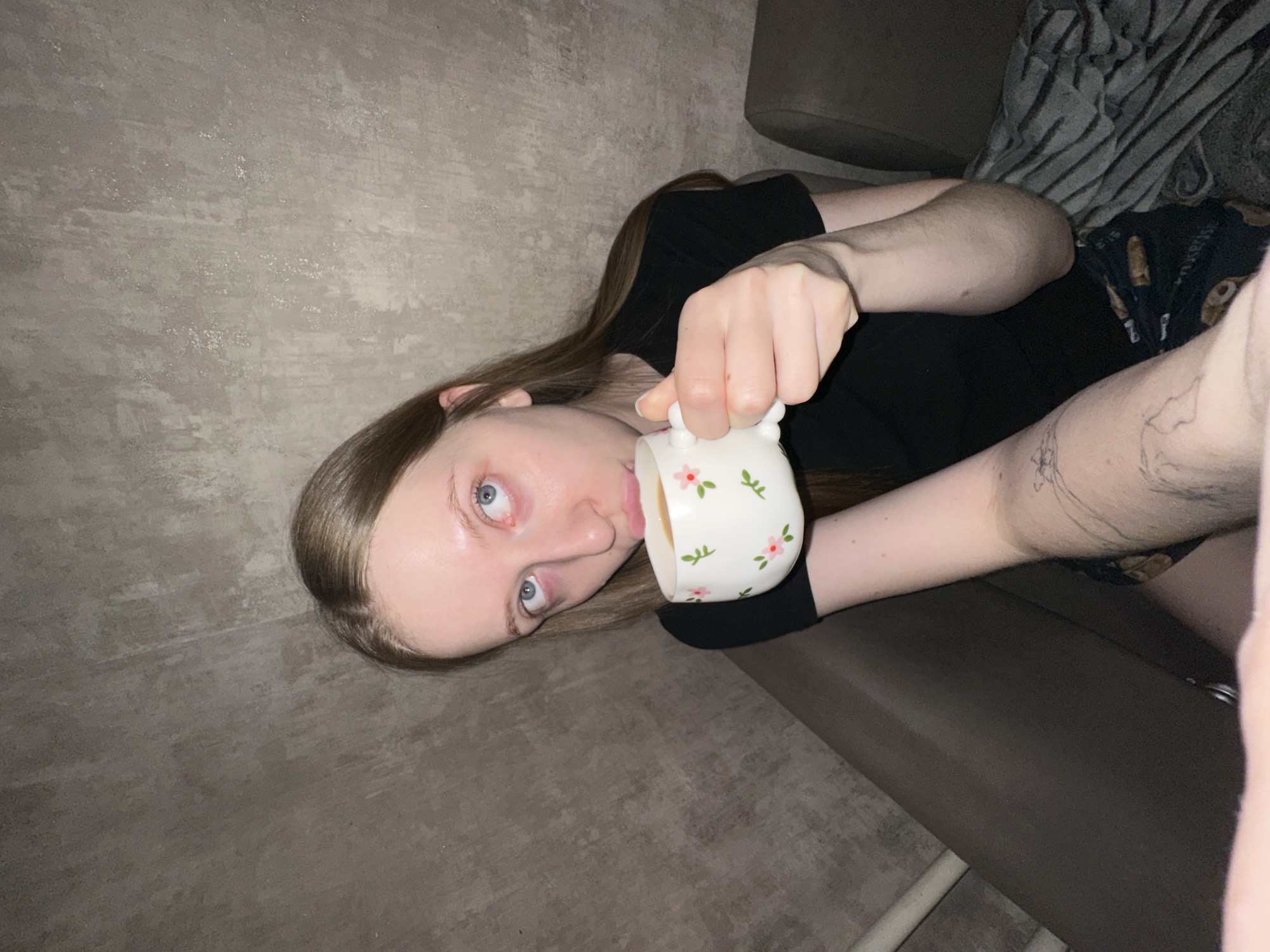 A young woman is lying on a grey couch and drinking from a white teacup decorated with pink and green flowers. She has long brown hair, pale skin, and blue eyes. She is wearing a black T-shirt and patterned shorts. There is a grey wall behind her, and a patterned blanket on the couch. The location is most likely indoors. There is no evidence in the image to suggest the location is in Chaikovsky, Russia.