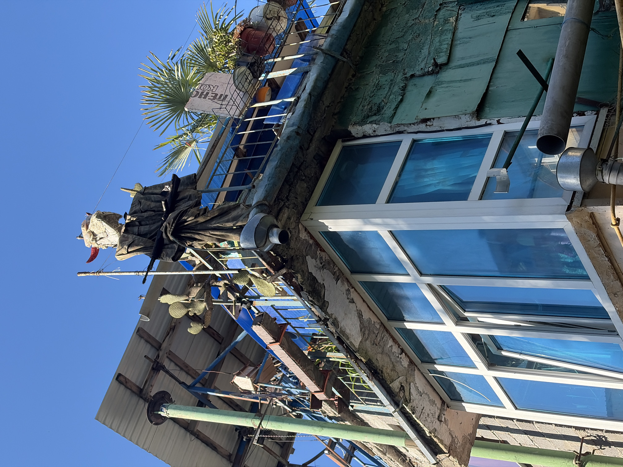 In a view looking up at a weathered, multi-story structure against a clear blue sky in Sochi, Russia, a peculiar scarecrow-like figure stands atop a balcony. The figure, dressed in dark, draped fabric, has a crude, mask-like head adorned with red horns. It appears to be holding what looks like a rifle. The building itself is a chaotic assembly of materials, featuring a corrugated metal roof supported by exposed wooden beams, blue plastic sheeting, and a metal railing. Cacti and potted plants are scattered on the upper levels. The lower portion of the building exhibits a large, modern window with bright blue-tinted glass, contrasting with the rough, crumbling concrete and peeling green paint of the surrounding walls. Various pipes and external conduits are visible, indicating a utilitarian and somewhat dilapidated state of construction. The overall impression is of an unconventional, possibly improvised, dwelling or structure, characteristic of some informal settlements or artistic modifications found in urban environments.