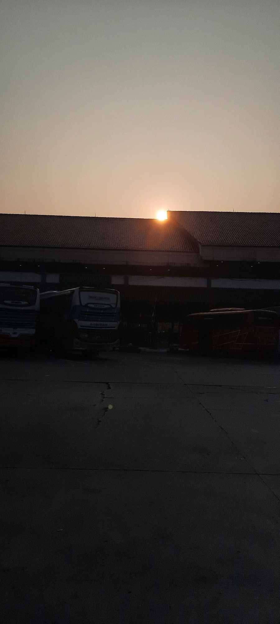 The image depicts a sunset in East Jakarta, Indonesia, likely at a bus terminal or a similar transportation hub. The sun is partially obscured by the roof of a large building, casting a warm, orange glow across the sky. Silhouettes of several buses are visible in the foreground, parked on a dark asphalt surface. The building appears to be a functional structure, possibly a terminal or a maintenance shed, with a dark tiled roof and some exposed structural elements beneath. The ground is cracked and weathered, suggesting an outdoor, utilitarian area. No people are clearly visible in the image. The overall atmosphere is one of quiet twilight, with the fading sunlight creating dramatic shadows.
