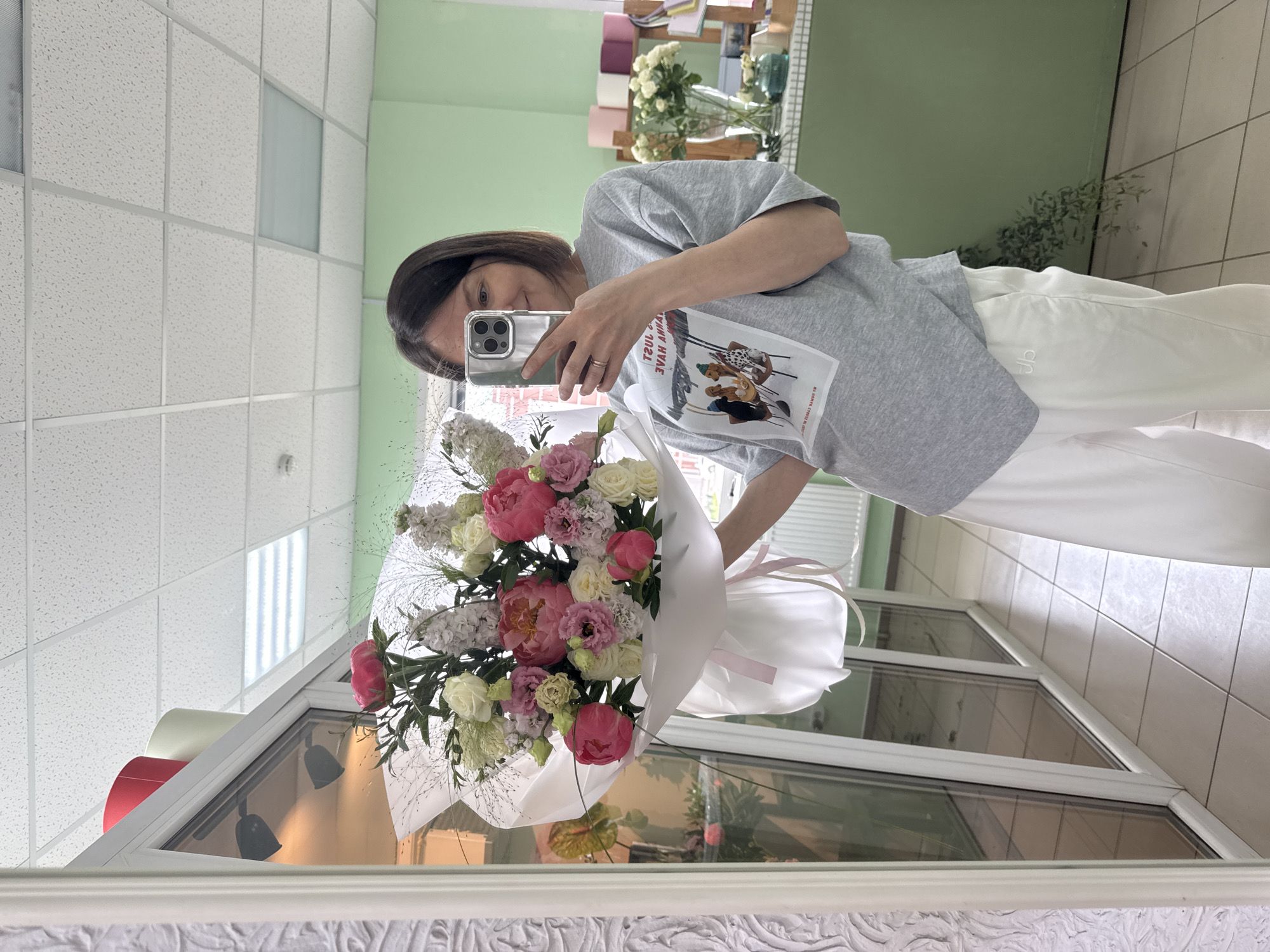 A young woman with dark hair and a grey t-shirt is holding a large bouquet of pink and white flowers in front of a mirror. She is taking a selfie with her phone, which is visible in her right hand. The bouquet is wrapped in white paper and tied with a pink ribbon. In the background, there is a wall painted light green, a shelf with various flower pots and vases, and a window. The floor is tiled with light beige tiles. The scene appears to be inside a flower shop.