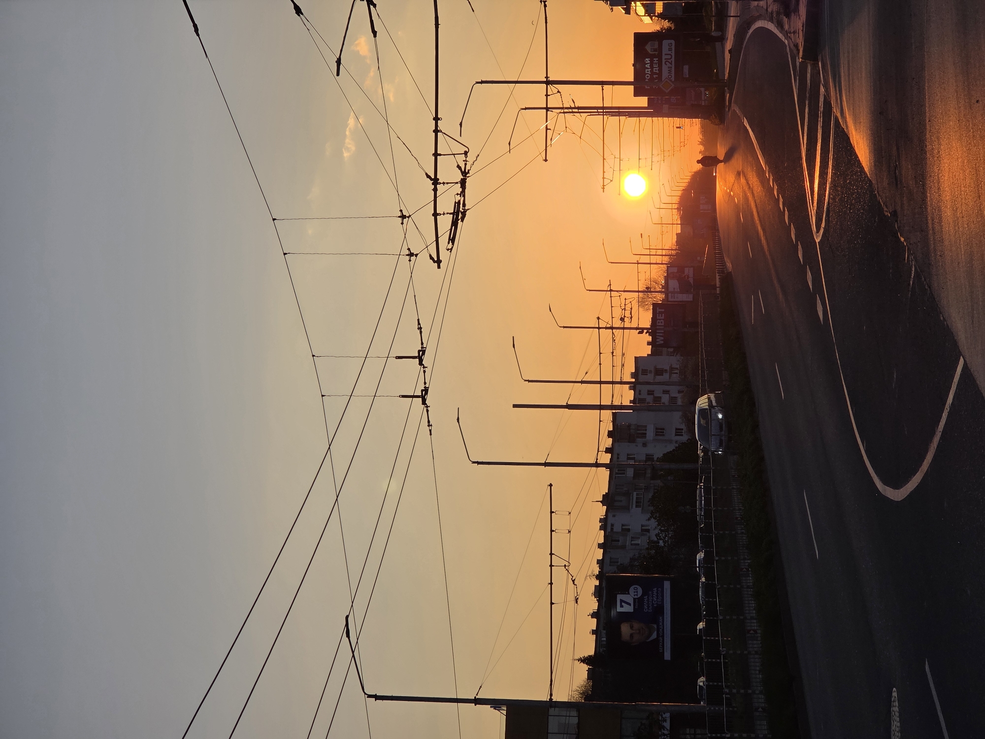 The image depicts a street in Varna, Bulgaria, at sunset. The sun is positioned in the center of the frame, casting an orange glow along the road. The road appears wet, reflecting the sunlight. Overhead tram lines and street lights are visible, silhouetted against the sky. Buildings line the street, along with political billboards. A car is visible on the road and there is a vehicle directly in front of the sun.