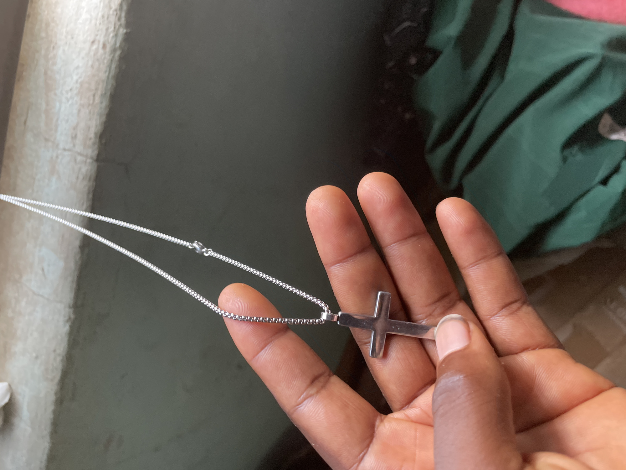 A close-up, overhead shot shows a person's hand holding a silver cross necklace. The hand is dark-skinned with short fingernails and is positioned in the lower left quadrant of the frame. The cross, a simple, unadorned design, hangs from a thin, silver chain that drapes across the fingers. The background is out of focus, with a light-colored, textured surface in the upper portion and a dark green fabric in the lower right. The lighting appears natural, casting soft shadows. This image was captured in Jalingo, Nigeria.