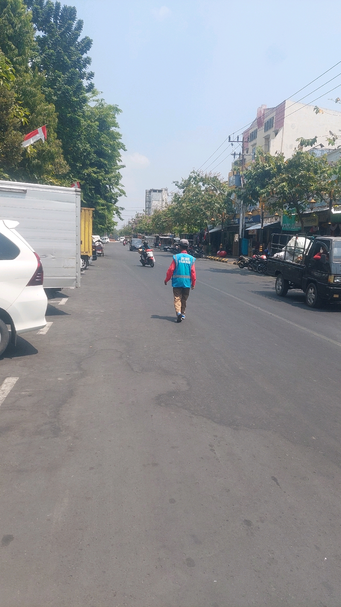 A street scene in Madiun, Indonesia, under a clear blue sky, features a man wearing a blue vest over a red shirt, walking away from the camera on the right side of the road. The vest has text printed on it, although it is not fully legible. He is also wearing brown pants and blue and black sneakers. To his left, a black pickup truck is parked. Further down the road, several motorcycles are visible, some parked and others in motion. On the left side of the road, a white car is parked alongside a large white truck. Trees line the street, and buildings with shops are visible on the right side. The Indonesian flag is displayed on a pole among the trees on the left. The road is asphalt, with white markings indicating parking spaces.
