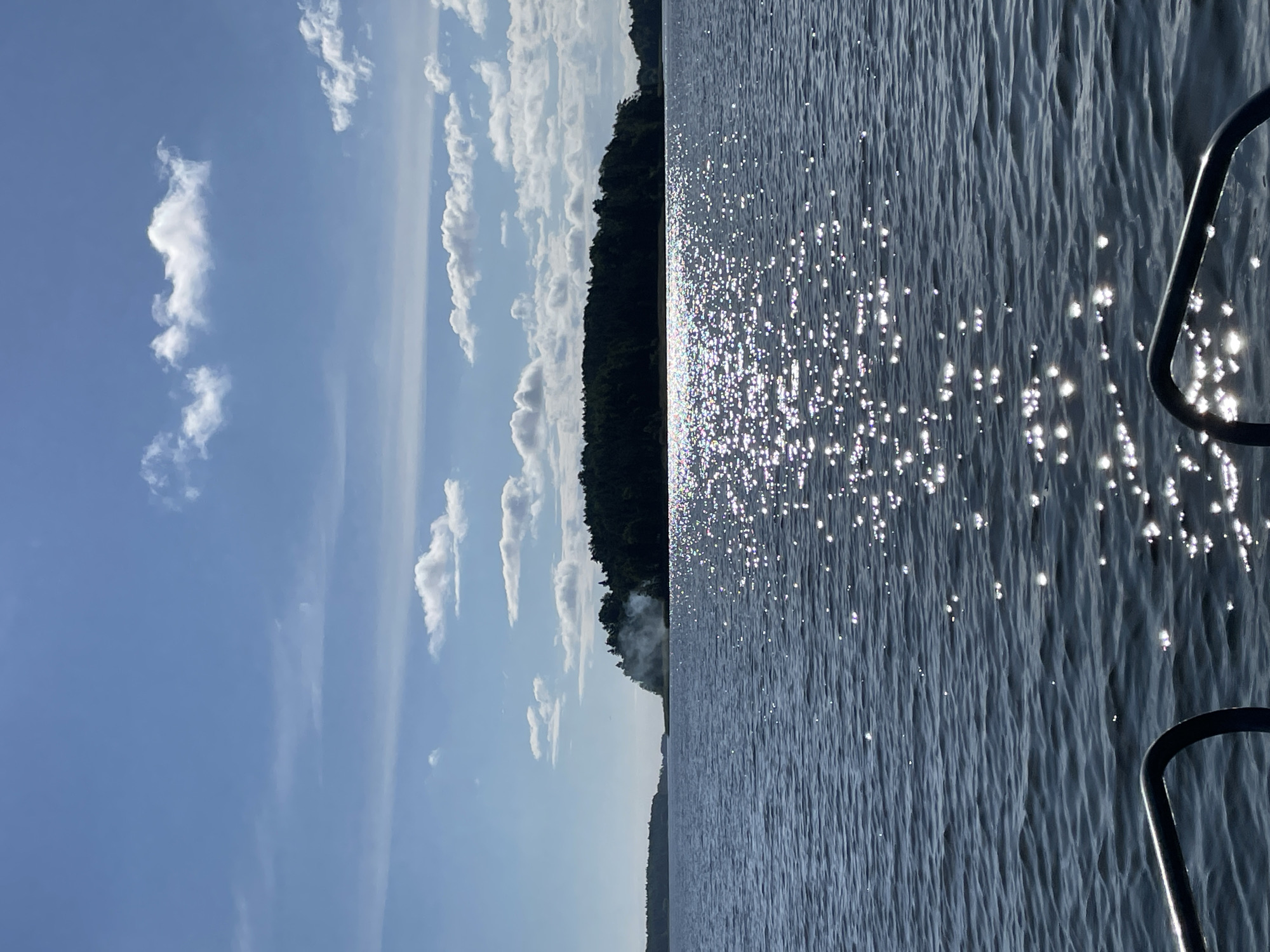 The image depicts a body of water, likely a lake or river, with a wooded island or shoreline in the distance. The sun is shining, creating a shimmering effect on the water's surface. White, puffy clouds are scattered across a clear blue sky. On the distant landmass, a plume of white smoke rises, suggesting a fire or other activity. The foreground shows a dark railing, possibly from a boat, suggesting the photo was taken from the water. The city of Berozovo, Russia, is a small town located in the Tyumen Oblast, known for its forests and proximity to water bodies. While the specific location within Berozovo is not identifiable from the image alone, the scene is consistent with the natural environment found in that region.