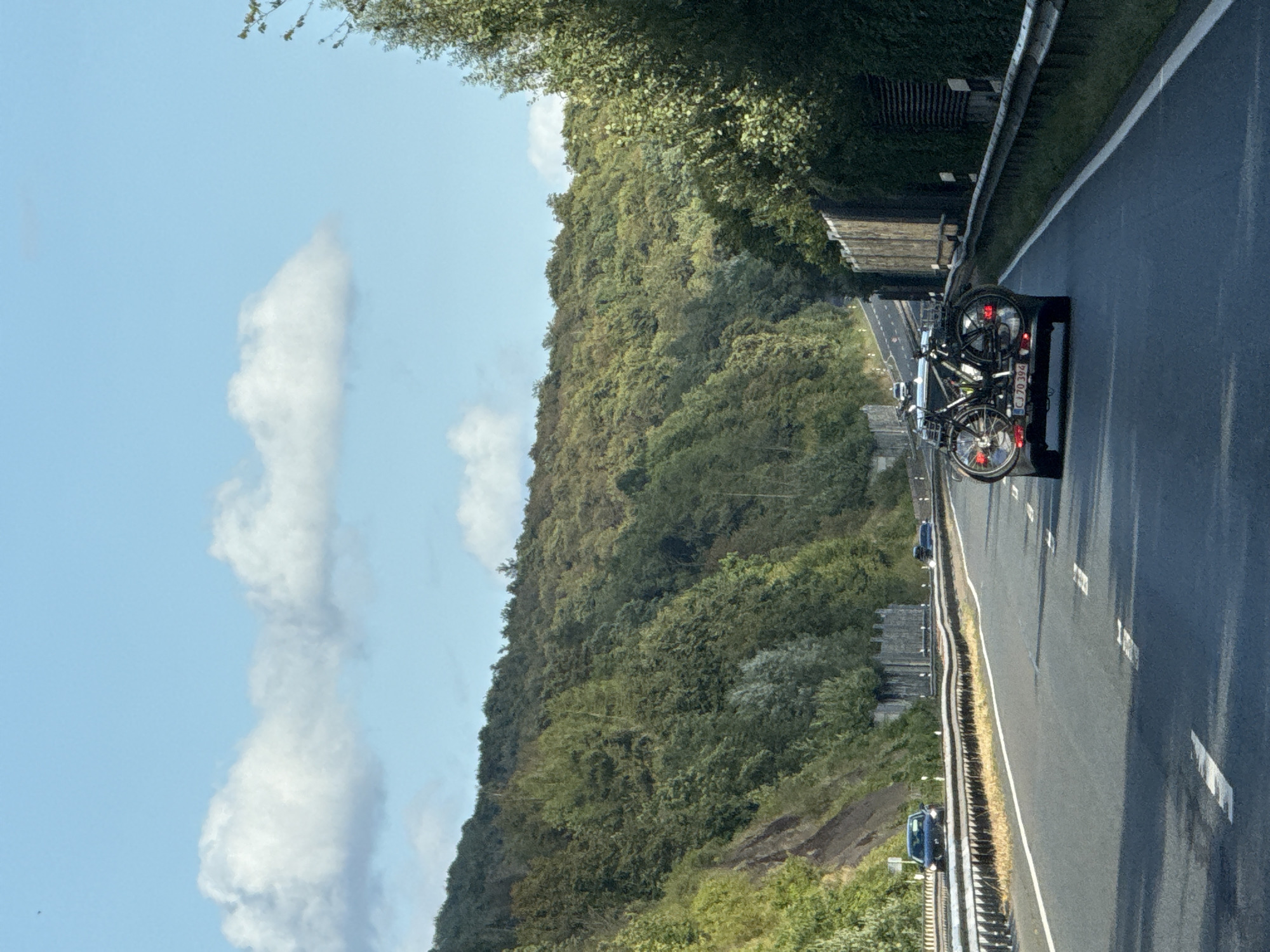 The image captures a view from a vehicle traveling on a highway in Herzberg, Germany. The highway is flanked by a lush, green, tree-covered hillside on the left and a dense line of trees and a sound barrier on the right. The sky is a clear blue with scattered white clouds. In the foreground, a dark-colored car is visible with two bicycles mounted on a rack on its rear. The car is in motion on the highway, as are other vehicles in the distance. The license plate on the car is partially visible and reads "CJ 70 394". The scene appears to be during daylight.