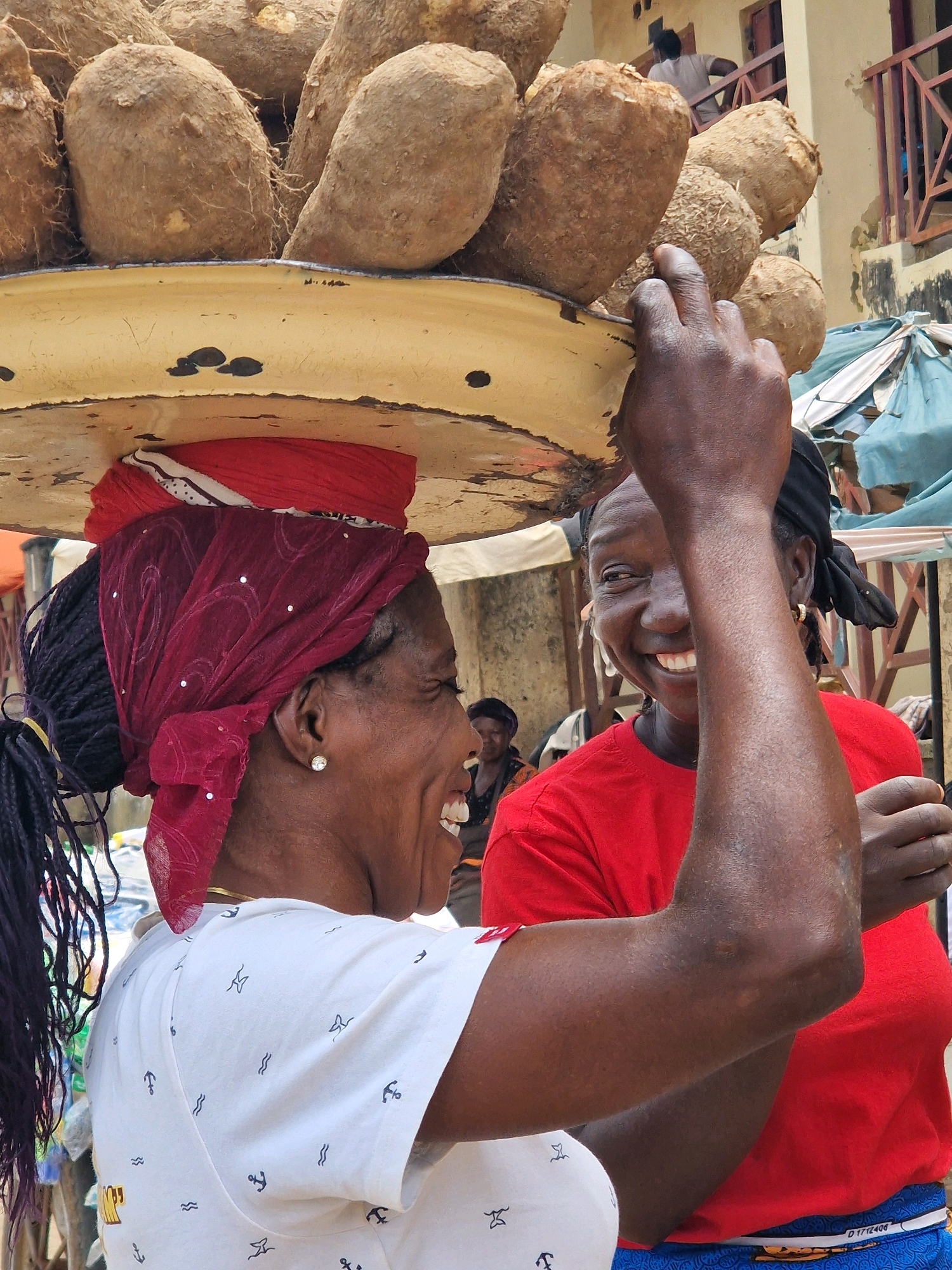 In an outdoor market setting, a woman with dark skin and dark hair, worn in braids, is balancing a large, round, light-colored tray on her head, laden with a substantial quantity of large, brown, root-like vegetables, possibly yams. She is wearing a red headscarf over a darker red fabric, both adorned with white embellishments. A portion of a red t-shirt is visible beneath the tray. To her right, another woman, also with dark skin, is looking towards the camera with a wide smile, her arm raised in front of her face. She is wearing a red t-shirt. In the background, a building with a light-colored facade and wooden railings is visible, as well as other market stalls and people. The overall scene suggests a vibrant market atmosphere, with the woman carrying the yams appearing to be either a vendor or a customer. The lighting suggests it is daytime.