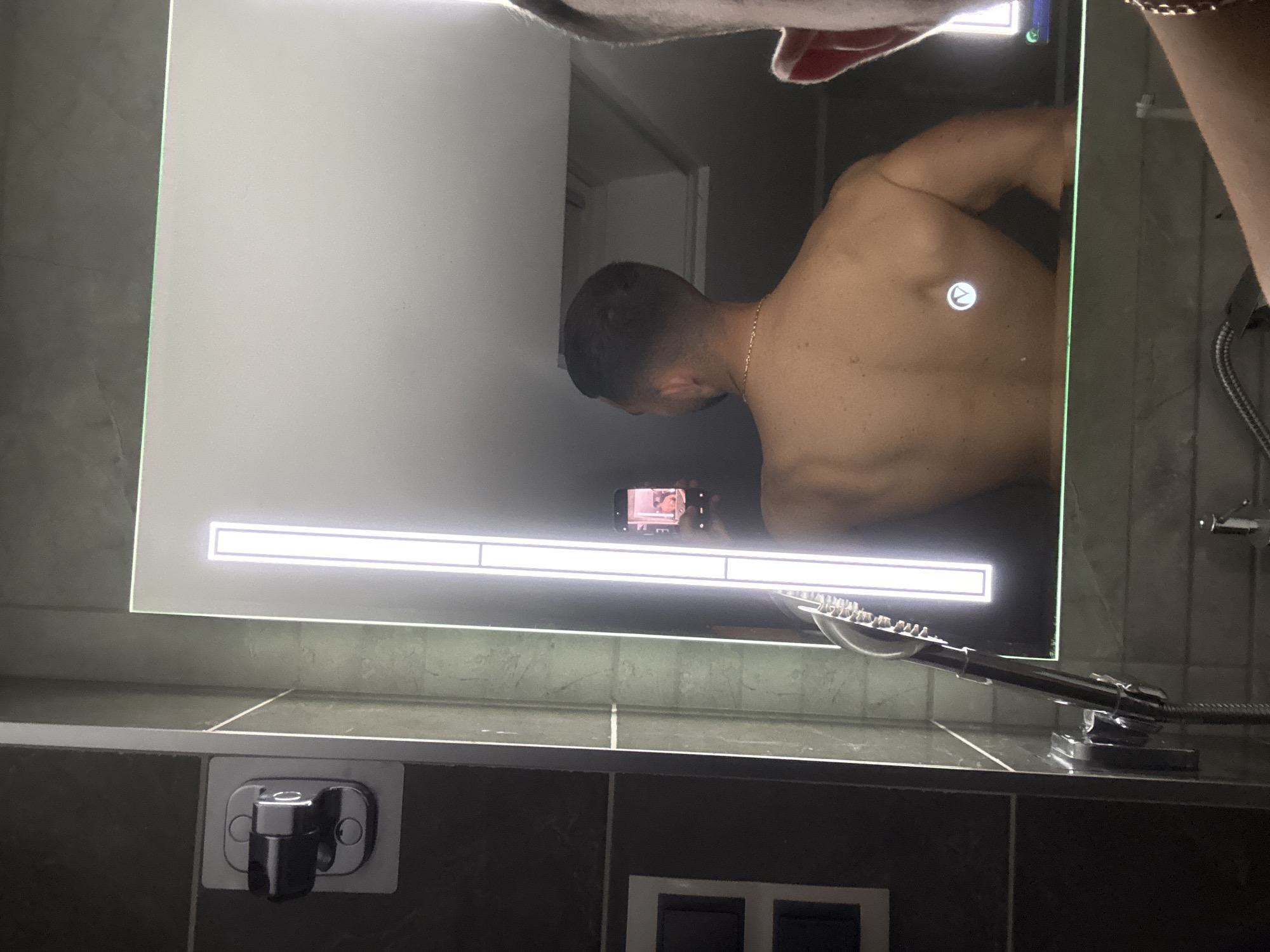 A person is taking a selfie in a bathroom mirror. The person's bare back and arm are visible in the mirror's reflection, along with their head and a gold chain. The person is holding a smartphone. Below the mirror is a countertop, tiled with dark brown and grey tiles. A toothbrush holder and a double light switch are also visible. The bathroom walls are grey. It is not possible to determine the location in the city of Gabrovo, Bulgaria from the image.