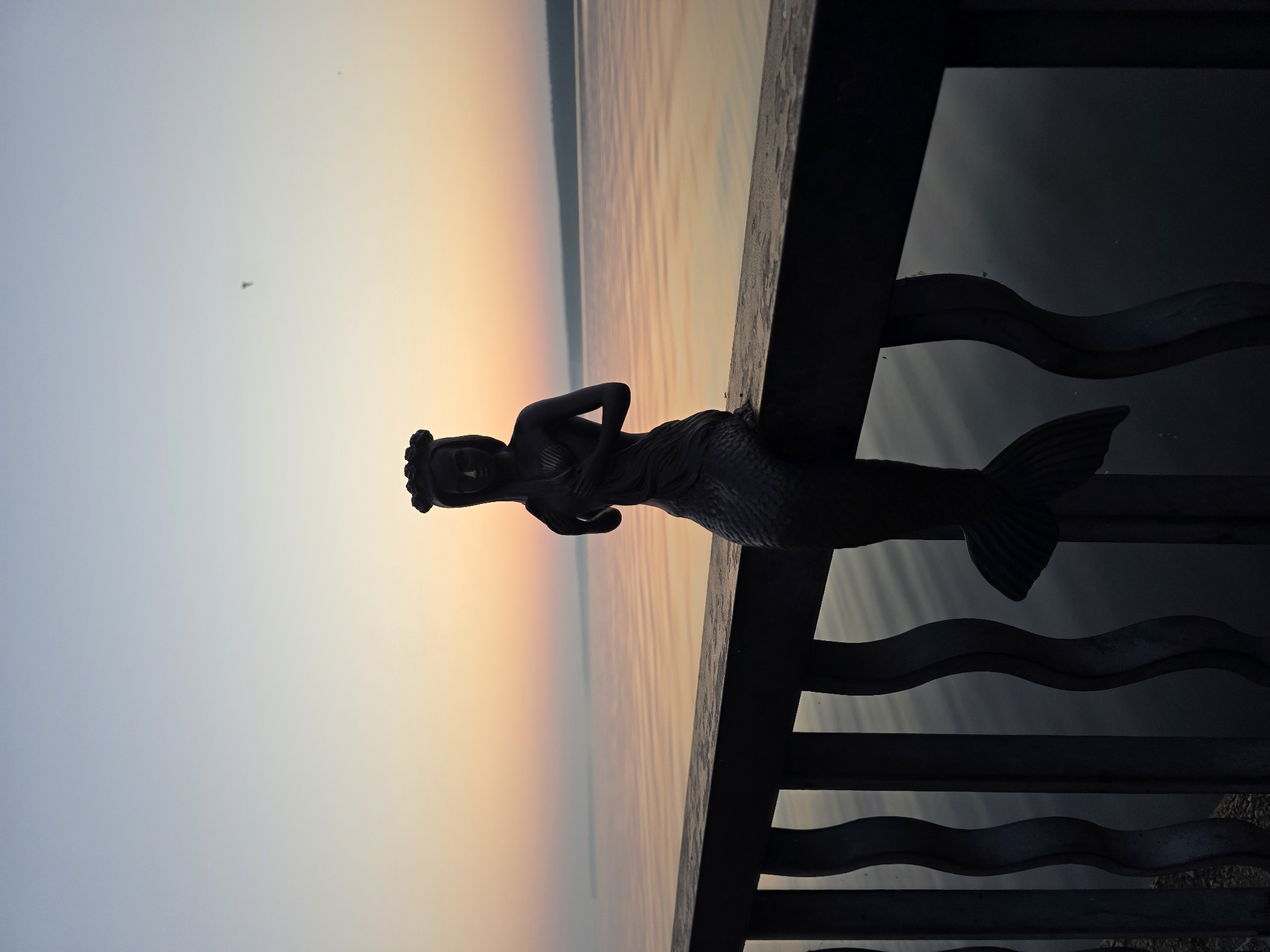 A dark silhouette of a mermaid statue sits on a dark metal railing overlooking a calm body of water at sunrise or sunset. The sky behind the water is a gradient of soft colors, transitioning from a light blue at the top to shades of orange and pink near the horizon. The water reflects these colors, creating a tranquil and serene atmosphere. The railing has an ornate, wave-like design, and a distant, hazy landmass can be seen on the horizon. The image appears to be taken from a viewpoint along the embankment of the Kama River in Chaikovsky, Russia.