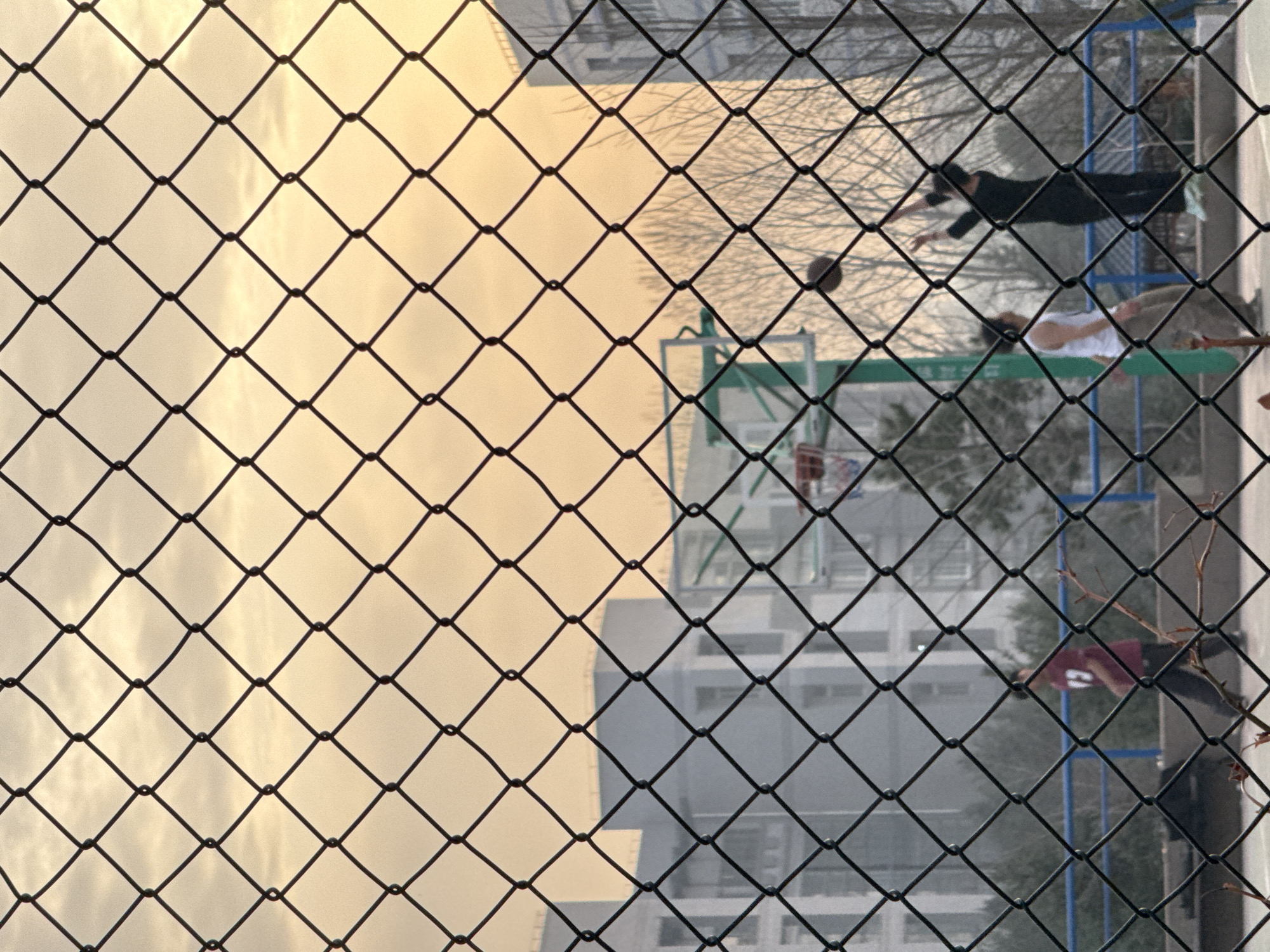 The image depicts a basketball court scene viewed through a wire fence. Three individuals are visible on the court. One person in a dark outfit is in the process of shooting a basketball towards the hoop. Another individual wearing a white shirt is positioned to observe the shot. A third person is standing at the side of the court, wearing a red shirt with the number "2" visible on the back. Behind the basketball court, there are buildings and trees. The sky is pale, and the light suggests that it might be either early morning or late afternoon. The general environment indicates a typical urban setting. The location is determined to be Zhongtun, China, based on the prompt.