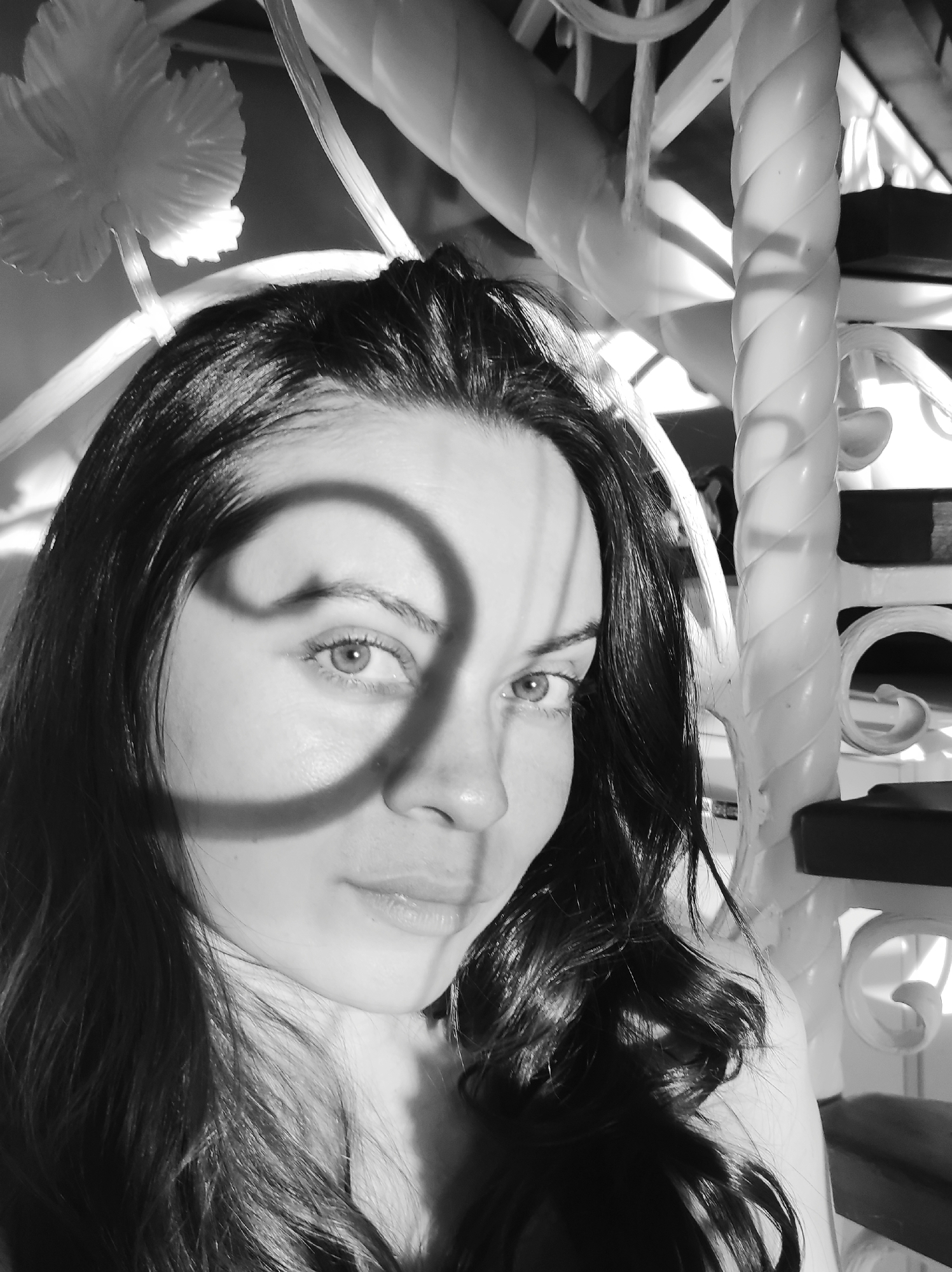 A black and white close-up photograph shows a woman with dark hair, lit with strong sunlight. The woman is facing the camera with a calm expression. An undefined shadow is cast on her face, originating from an unseen source. Behind the woman, partially obscured and out of focus, is a white decorative metal structure, appearing to be part of a railing or furniture with curving and spiraled components. The location is Unknown, Unknown.