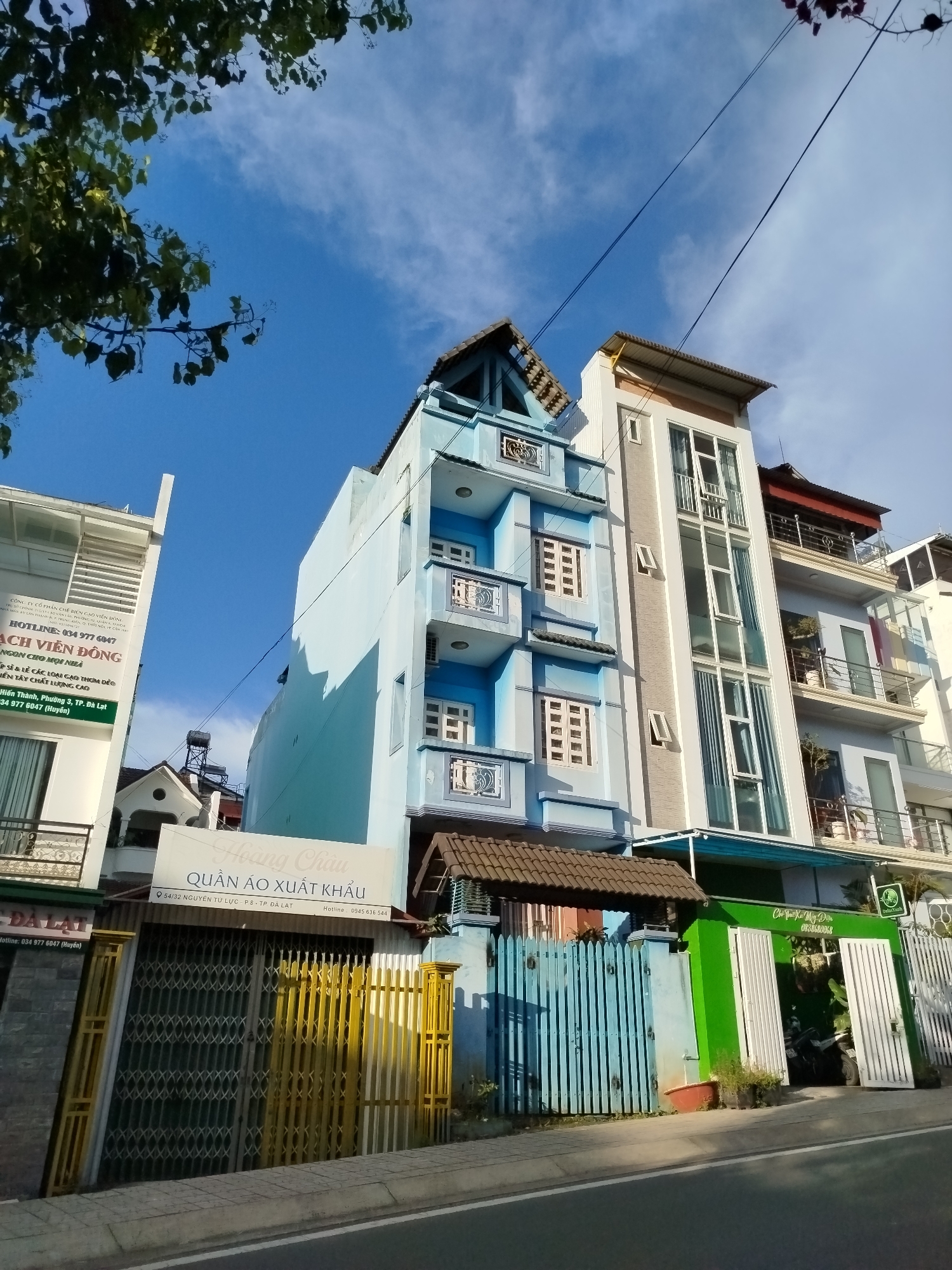 This image captures a street scene in Da Lat, Vietnam, featuring a row of multi-story buildings. The architecture is varied, with a prominent light blue building displaying ornate balconies and white-framed windows. Adjacent to it is a taller, modern white building with a facade dominated by large glass windows. Further down the street, other buildings with balconies and varying colors are visible.
In the foreground, a portion of a street is visible, with a sidewalk and a yellow-colored metal fence. There is also a green fence with white doors, and a blue wooden fence. Some vegetation can be seen near the fences. The sky above is clear and blue with scattered white clouds, and the top branches of a tree with green leaves are visible in the upper left corner.
Signage on the buildings indicates businesses. One sign, in Vietnamese, reads "HOÀNG CHÂU QUẦN ÁO XUẤT KHẨU," which translates to "Hoang Chau Export Garments." Another sign on a white building also appears to be for a business with contact information. The overall impression is a lively street with commercial activity in a Vietnamese city.