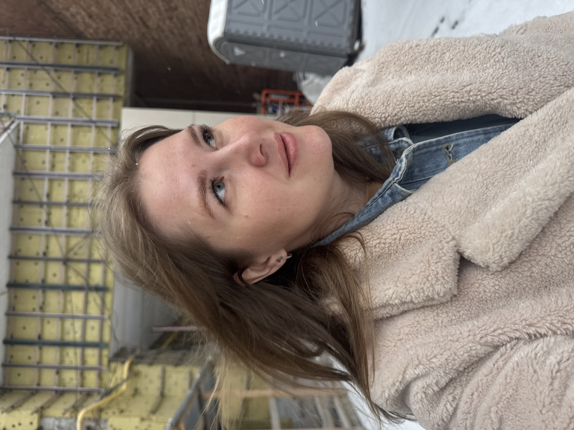 A close-up, vertical portrait captures a young woman outdoors in Perm, Russia, against a blurry industrial or construction backdrop. The woman is positioned from the chest up, looking upwards and slightly to her right with a serene, contemplative expression. She has light brown hair and blue eyes. She is wearing a light beige, fluffy, teddy-bear-style coat over a blue denim jacket. The background on the left features a structure with yellow insulated panels and a metal grid framework. To the upper right, a grey portable toilet-like unit is visible. Snow covers the ground in the background, indicating cold weather conditions. The scene is illuminated by daylight. No text is visible in the image.