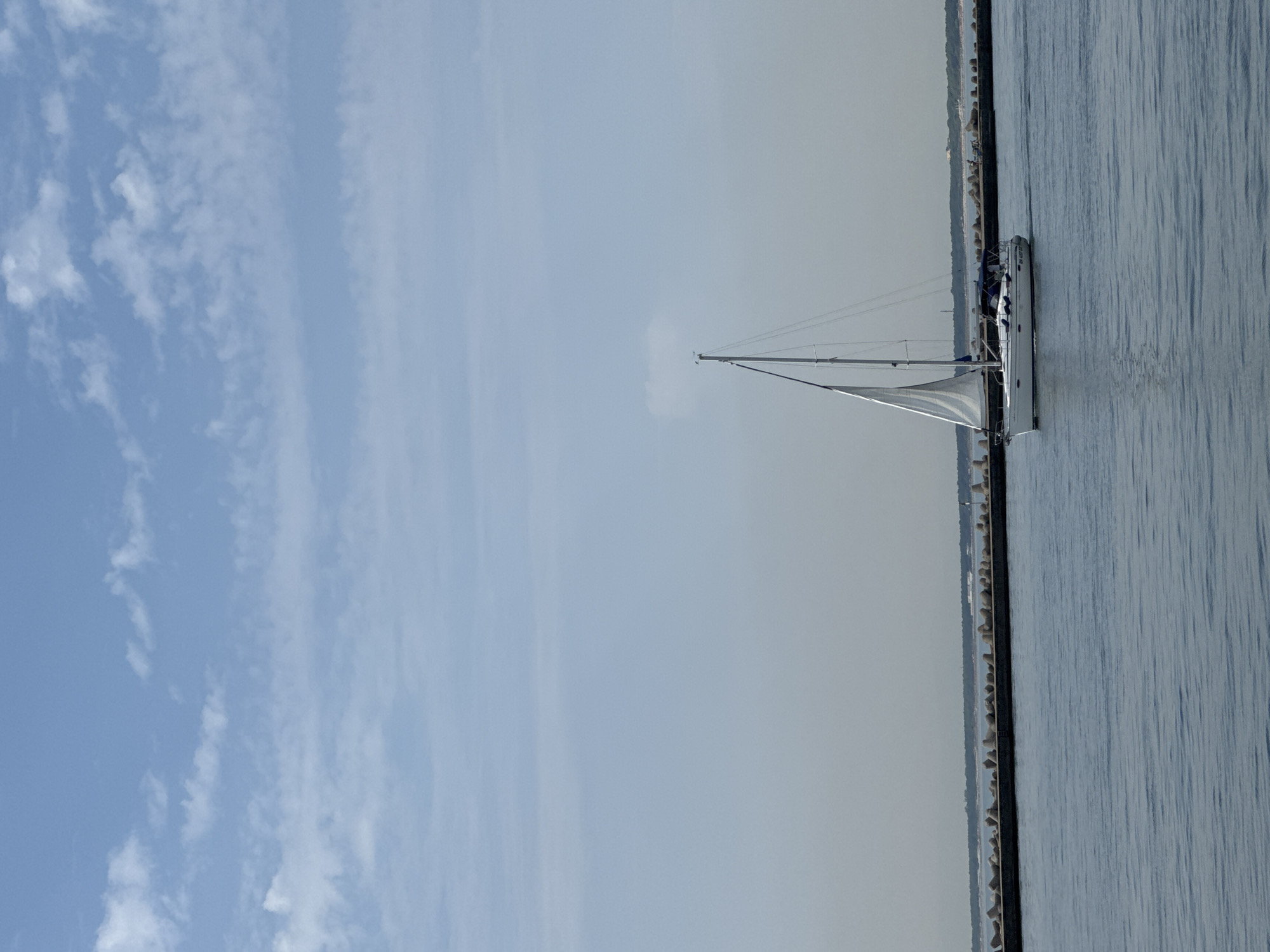 A sailboat with white sails is moving across blue water. In the background, there is a long breakwater made of concrete blocks and a distant shoreline with trees and buildings. The sky above is light blue with scattered white clouds. The scene appears to be a harbor or coastal area. There is no discernible connection to Ravenna, Italy, in the image.