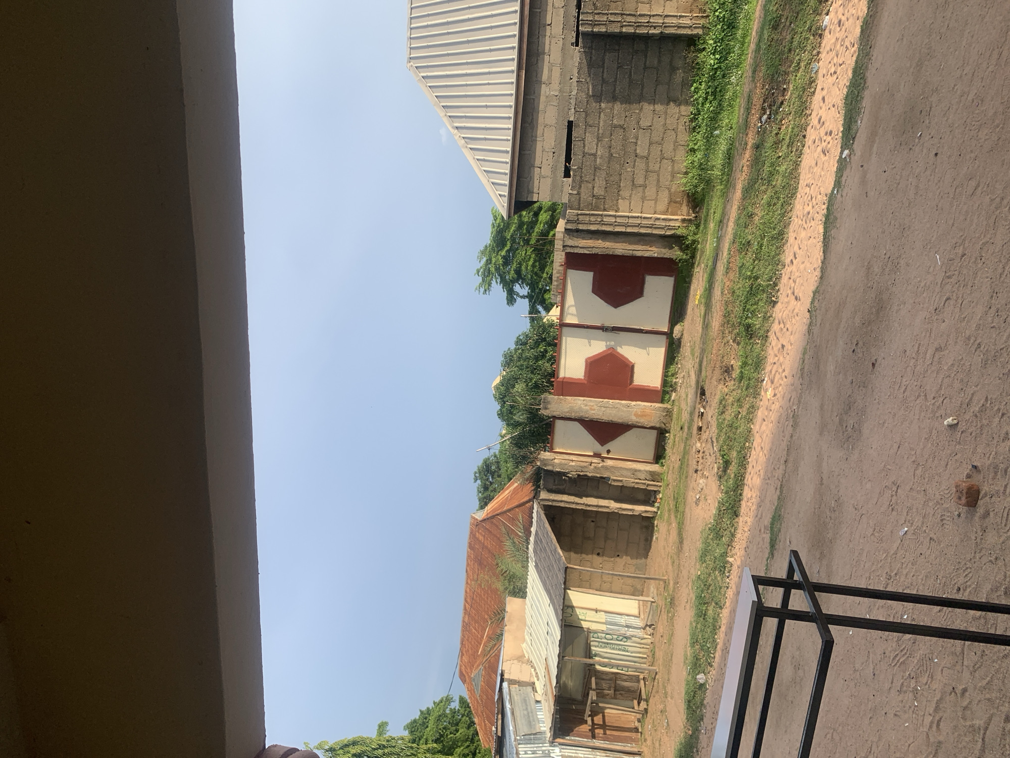 The image depicts an outdoor scene, likely in Jauro Yinu, Nigeria. The perspective is low and to the side, possibly from under a covered structure indicated by a large, dark, out-of-focus object on the left, which could be part of a building's overhang or eaves.
The background shows a street or dirt path running diagonally from the foreground to the middle ground. Along the right side of the path is dry, sandy earth with some scattered debris. To the left of the path, there's a strip of green grass, then a low wall or fence made of concrete blocks. Beyond this wall, a two-part gate stands, painted with red and white diamond shapes. Further back, there are various buildings. One has a pitched roof made of corrugated metal, which appears rusted in places. Another building to its right is constructed from concrete blocks and also has a metal roof. Trees with lush green foliage are visible in the background, peeking out from behind the structures. The sky is clear and light blue, suggesting it is daytime. The lighting indicates it could be morning or afternoon. There is no discernible activity or people in the image. The overall impression is of a rural or semi-urban environment in Nigeria, with a mix of simple construction and natural elements.
