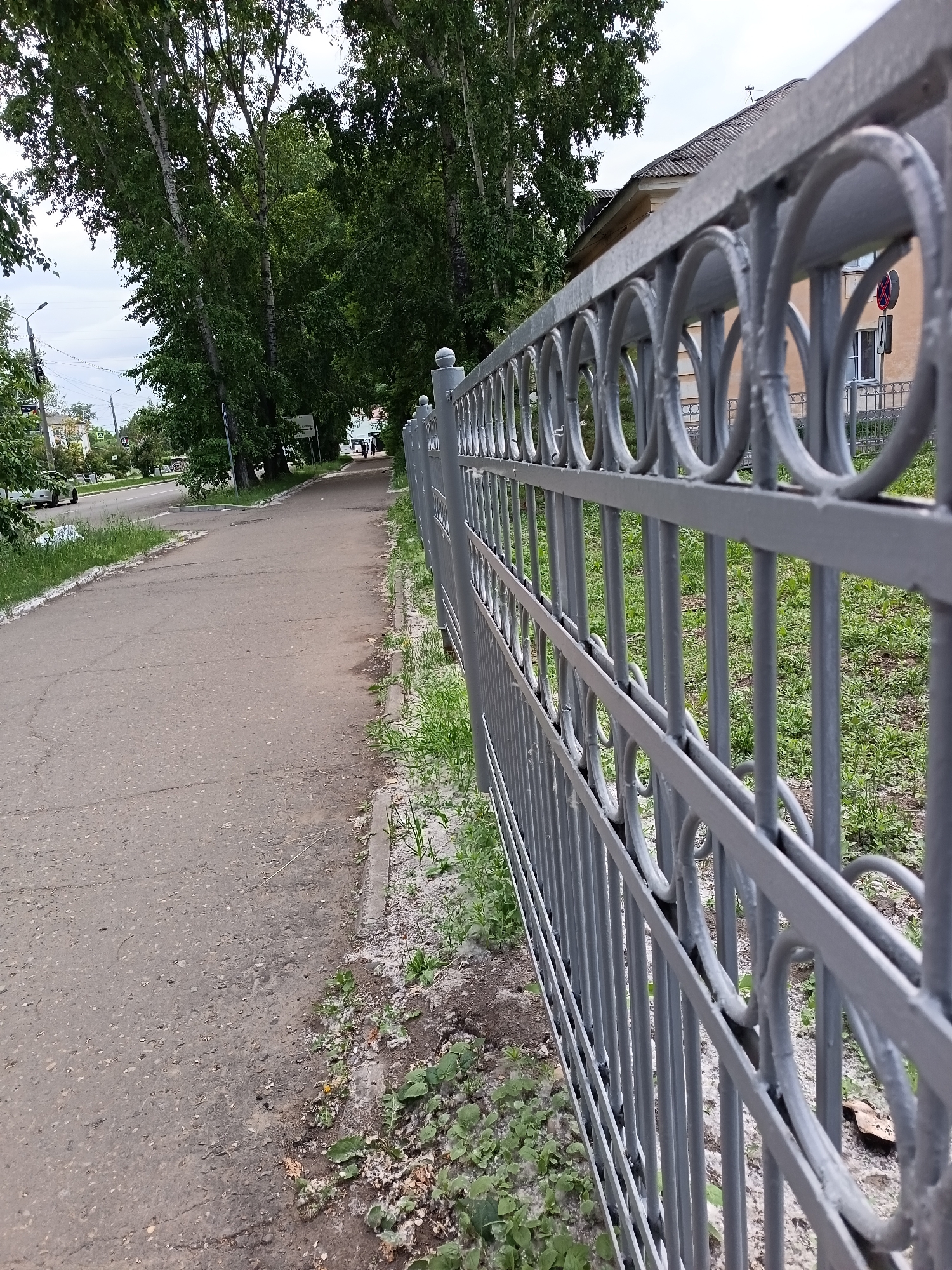 The image shows a grey, ornamental metal fence bordering a paved pedestrian walkway. The walkway is to the left of the fence and is cracked and weathered, with a strip of grass and dirt separating it from the fence. Beyond the walkway, a road is visible with some trees lining it and buildings in the distance. A white car is parked on the road. The fence has vertical bars and a decorative element consisting of circles with smaller circles inside. The fence is the primary subject of the foreground, with the walkway and street receding into the background. The overall scene depicts an urban or semi-urban environment in Komsomolsk-on-Amur, Russia, likely during the daytime with overcast skies.