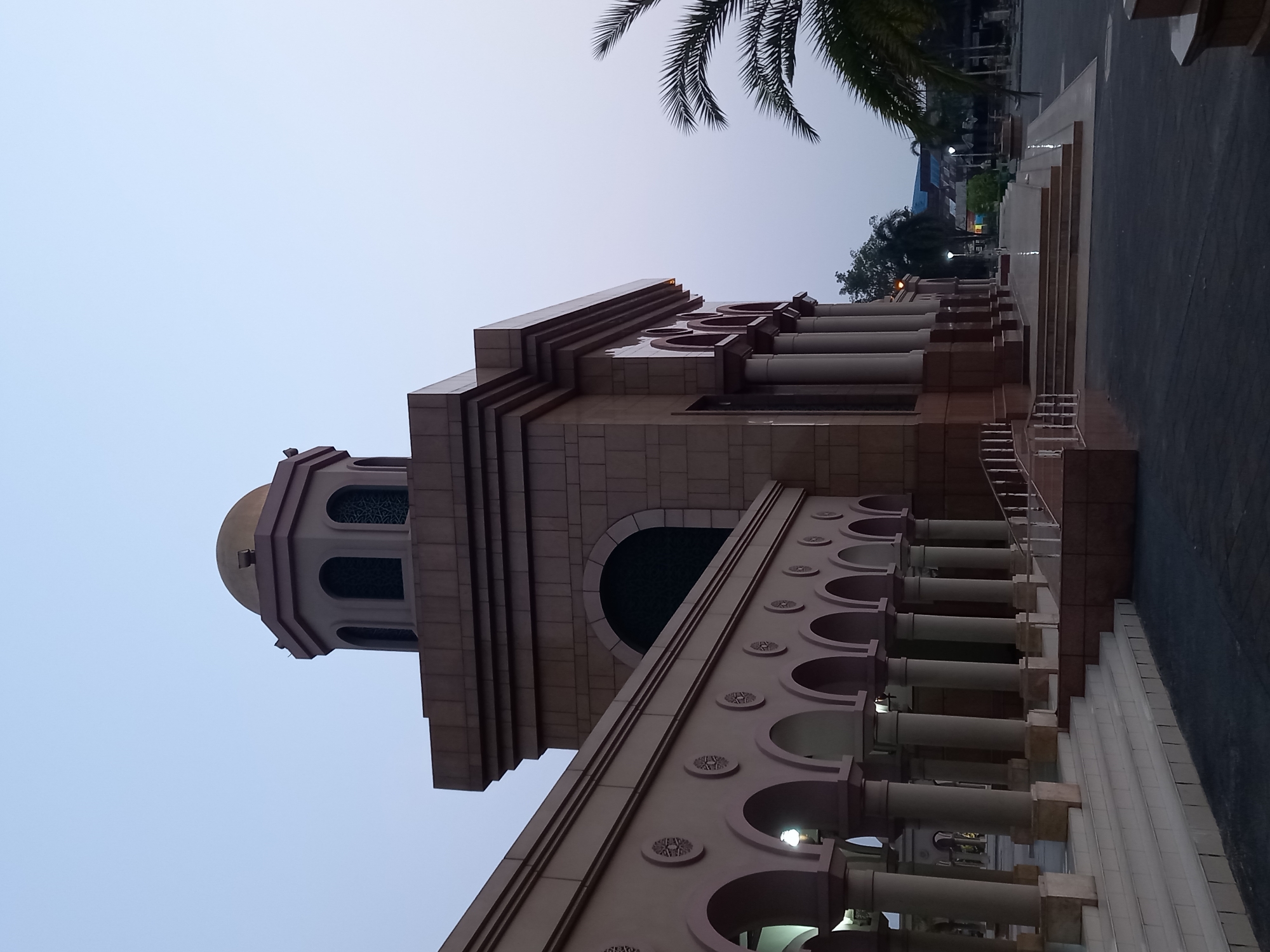 The image shows the exterior of the Islamic Center Samarinda, also known as Masjid Raya Samarinda, located in the city of Samarinda, East Kalimantan, Indonesia. The building features a prominent golden dome and a tower with arched windows. A long, colonnaded walkway with a series of arches and decorative circular elements extends from the main structure. The architecture is characterized by its stone facade, classical columns, and elaborate detailing. Steps lead up to the entrance. In the foreground, there is a paved area and to the right, the fronds of a palm tree are visible. The sky is a pale, clear blue, suggesting it is either early morning or late afternoon. No people are present in the image. The surrounding environment appears to be a public space adjacent to the mosque.
