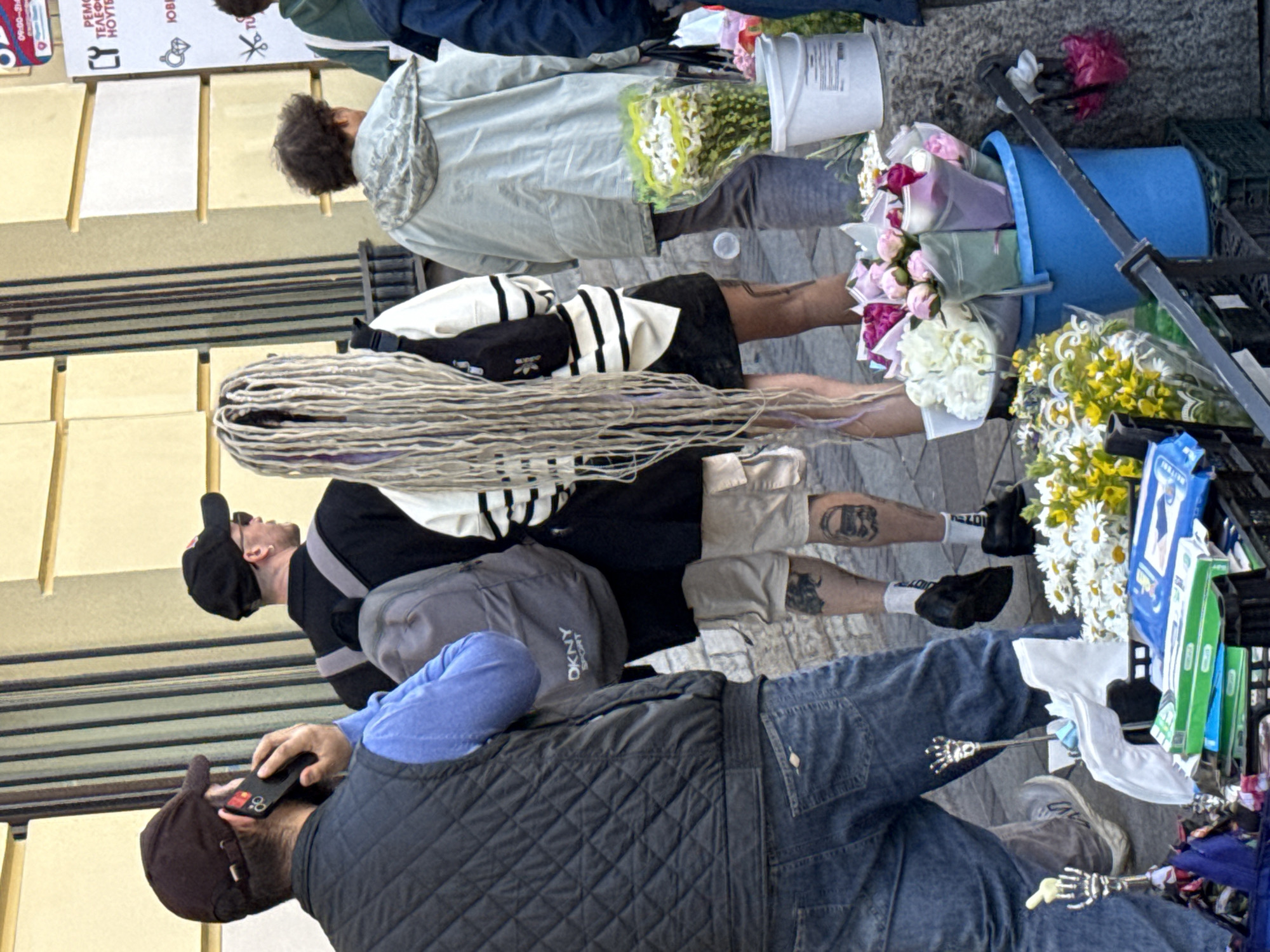 The image depicts a street scene in Saint Petersburg, Russia, likely during the daytime with fair weather, as indicated by the natural lighting and outdoor setting. The scene features several individuals, some of whom appear to be vendors or shoppers.
In the foreground, a man wearing a quilted vest and jeans is seen holding a smartphone to his ear. He has a baseball cap on. To his left, another person wearing a black baseball cap and a black jacket over a white and black striped shirt is visible. This individual has long, light-colored dreadlocks. Further back, a person in a light-colored jacket and dark pants is standing near a white bucket filled with flowers. Another individual is seen wearing a white and black striped jacket and beige shorts, with tattoos visible on their legs.
Objects in the scene include several buckets, one blue and one white, containing bouquets of flowers, predominantly pink, white, and yellow. There are also crates and what appear to be goods displayed on a street stall or table.
The overall impression is that of a bustling outdoor market or street-side vendor area. The presence of flowers suggests a possible flower market or a vendor selling bouquets. The clothing worn by the people is casual and suitable for outdoor activity. The location cues are consistent with an urban street environment. There is no discernible text in the provided image, other than a partial logo on a gray bag that appears to say "DKNY SPORT".