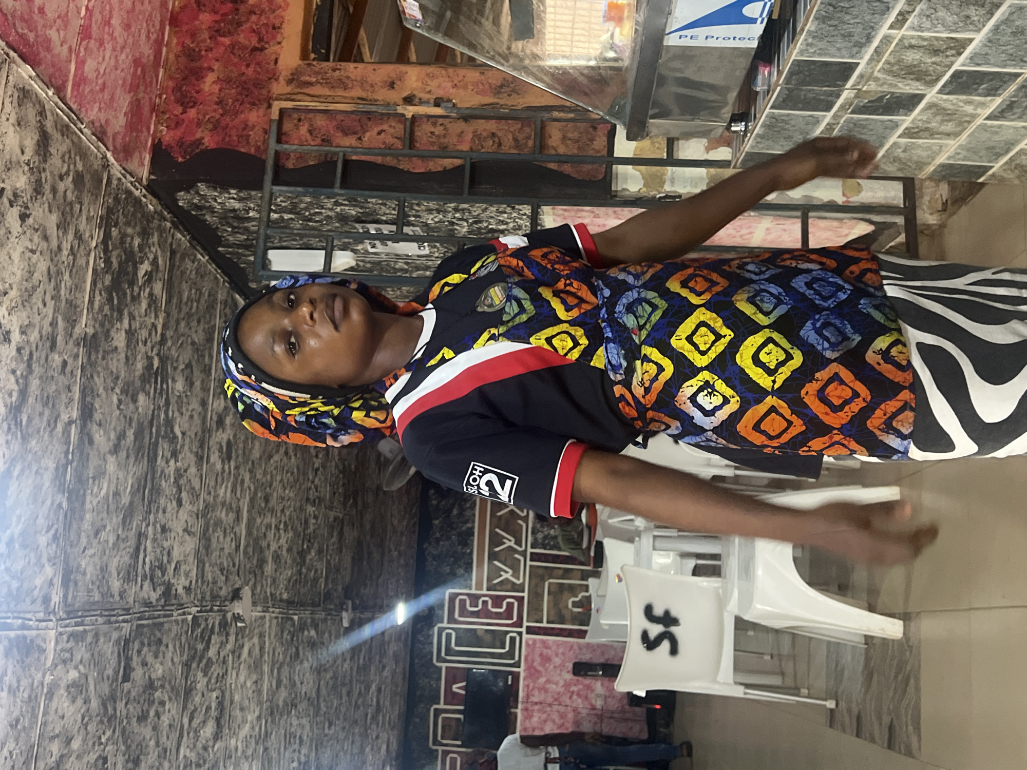 A Black woman with her hair covered by a colorful headscarf stands in what appears to be a restaurant or food stall in Nkpor, Nigeria. She is wearing a black t-shirt with red and white trim and a logo on the left sleeve. Over her shirt, she wears a vibrant, multi-colored patterned apron. Her lower body is clad in black and white striped pants. She is looking towards the viewer with a neutral expression.
The environment suggests a commercial setting. In the background, there are white plastic chairs and tables. A large, decorative sign with block letters spelling "PEACE" is visible on the wall behind the chairs. To the right of the woman, a glass display case, possibly for food, is present. The walls and ceiling have a textured, possibly industrial or rustic finish, with exposed pipes and wires visible on the ceiling. The overall lighting is somewhat dim, with a bright light source illuminating the area from the left.