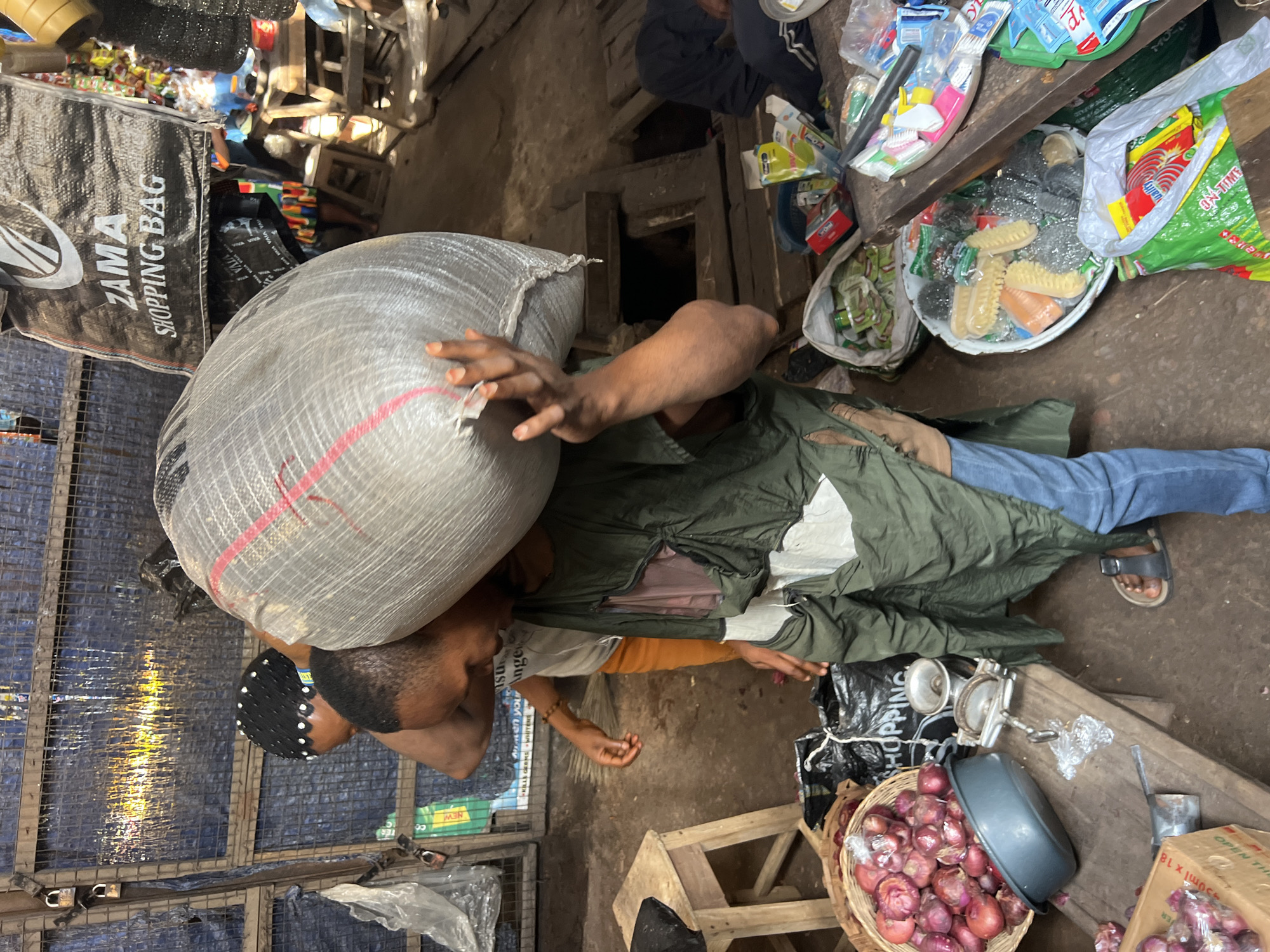 In Nkpor, Nigeria, a market scene unfolds with a person carrying a large, grey, netted bag filled with unknown contents over their shoulder. The bag has red and pink stripes running vertically. The individual is wearing a green garment over a tan-colored shirt and blue jeans, with black sandals. To the left, another person with a black, beaded headscarf is partially visible, reaching out. In the foreground, a basket overflows with red onions, next to a gray basin. A small metal grinder sits on a wooden bench, surrounded by plastic bags and various items, including cleaning supplies like brushes and sponges, and packaged goods. The background shows a rustic structure with metal fencing and a blue tarp.