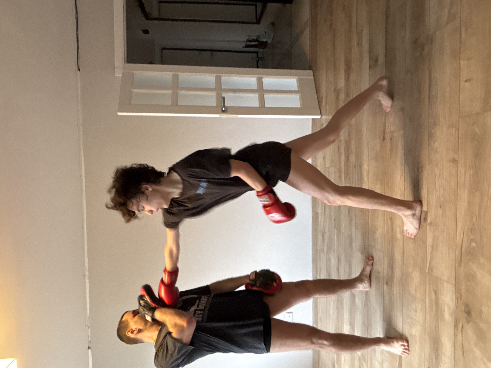 Two people are in a room with light brown wood flooring and white walls. A partially opened white door with multiple square panes is in the background. One person is standing closer to the door, wearing a dark t-shirt and dark shorts. They are wearing red boxing gloves. They appear to be throwing a punch downward. The second person is closer to the viewer, also wearing a dark t-shirt and shorts. They are wearing red and black boxing gloves and are partially covering their face with one of the gloves. The image does not allow determination of the location being Chaikovsky, Russia.