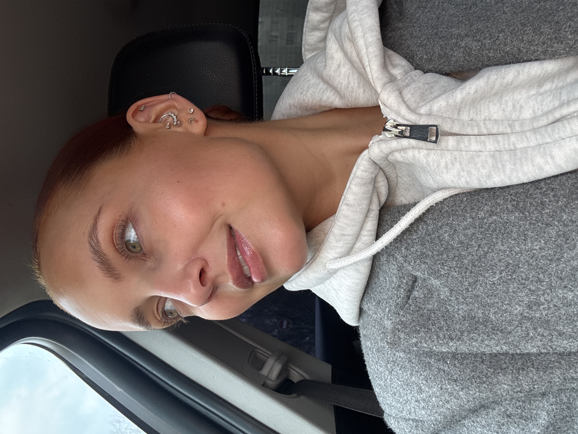 A young woman is captured in a close-up, upward-angled portrait, likely a selfie, inside what appears to be a car. She has light brown or auburn hair pulled back, and striking green or hazel eyes that gaze directly at the viewer. Her lips are slightly parted, revealing a subtle smile and a hint of lip gloss.
She is wearing a light grey, flecked zippered hoodie, with the zipper pull visible on her chest. Her left ear is adorned with several piercings, including small hoops, studs, and a curved barbell, adding a distinctive detail. The lighting suggests an overcast or cloudy day, with a grey sky visible through the car window in the upper left background.
Behind her, a dark car seat headrest is partially visible. To her right, a dark grey, possibly fleece-covered car seat or fabric is seen. A black seatbelt strap is also visible in the lower right, confirming the indoor automotive setting. The overall mood is calm and casual. There is no visible text in the image. The context places this scene in Chaikovsky, Russia.