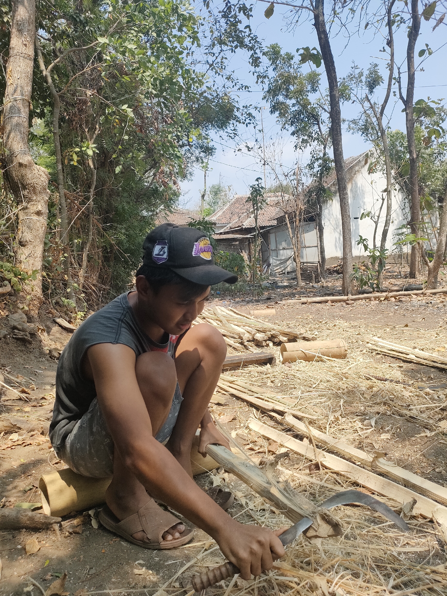 In Tongaswetan, Indonesia, a young male wearing a grey t-shirt, denim shorts, a black baseball cap, and sandals is crouching on the ground. He is actively working with a curved blade tool, shaping a piece of bamboo. The surrounding environment is rustic, with numerous bamboo stalks and dried leaves scattered across the dirt ground. In the background, traditional Indonesian houses with tiled roofs and white walls are visible, nestled amongst trees and foliage under a clear sky.