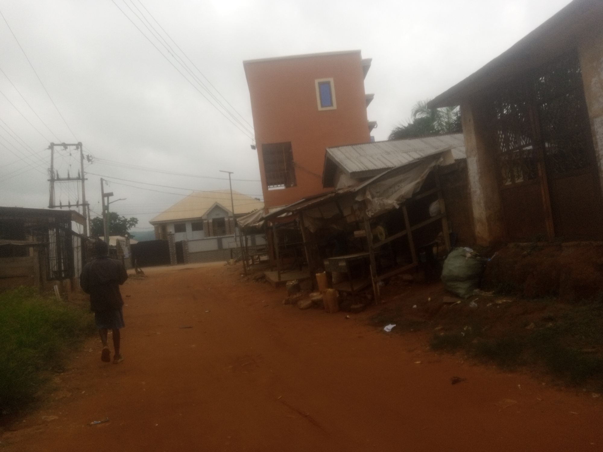 In Nanka, Nigeria, an individual walks along a dirt road lined with structures, including a multi-story orange building with a blue window and a tin-roofed market stall. Electrical poles and wires are visible in the background. The scene appears to be a commercial area with some debris and vegetation present.
