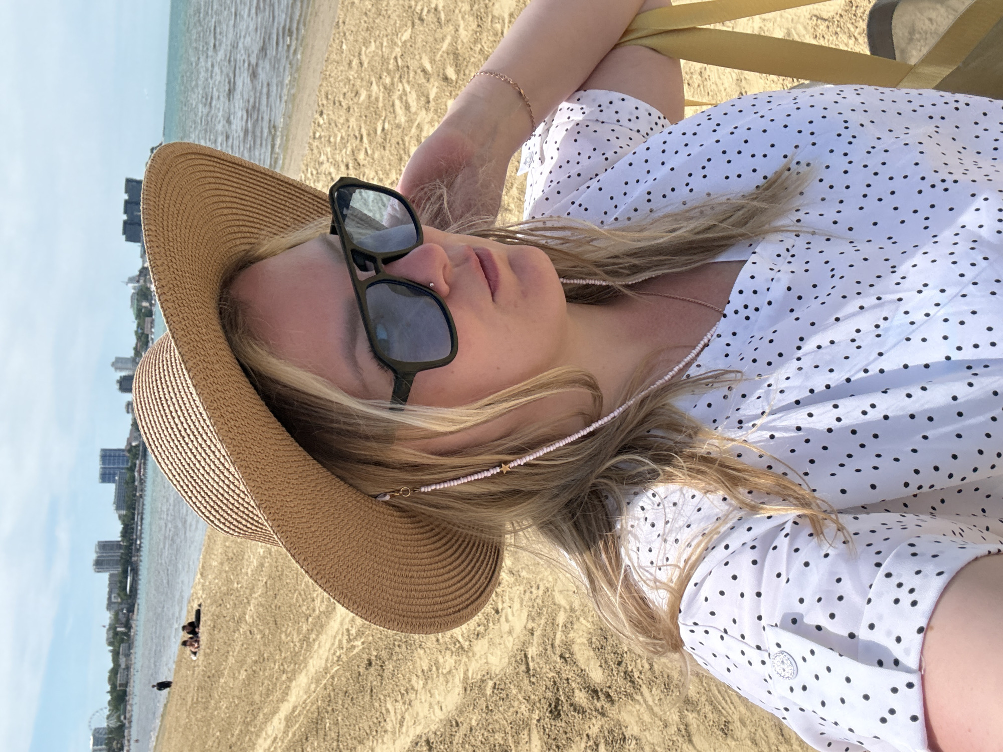 This image captures a close-up, slightly overhead view of a blonde woman relaxing on a sandy beach. She is wearing a wide-brimmed straw hat and dark-framed sunglasses, with a small nose piercing visible. Her long blonde hair is draped around her shoulders and across her chest. She is dressed in a white shirt with numerous small black polka dots. A delicate, light pink beaded chain with a small gold star charm adorns her neck, likely functioning as a glasses chain. A thin gold bracelet is visible on her left wrist. A yellow strap, possibly from a beach chair or bag, is also seen in the upper right.
The woman is looking upwards, suggesting she is reclining or lying down, enjoying the bright, sunny weather. In the background, a vast expanse of sandy beach meets the calm, light blue-green water of the sea. Along the distant coastline, a city skyline featuring multiple buildings is visible under a clear, bright sky. Small, indistinct figures of other people are scattered on the far beach, and a faint Ferris wheel can be spotted in the distance. The setting suggests a tranquil day at a coastal resort, specifically Anapa, Russia, conveying an atmosphere of relaxation and leisure. No visible text is present in the image.