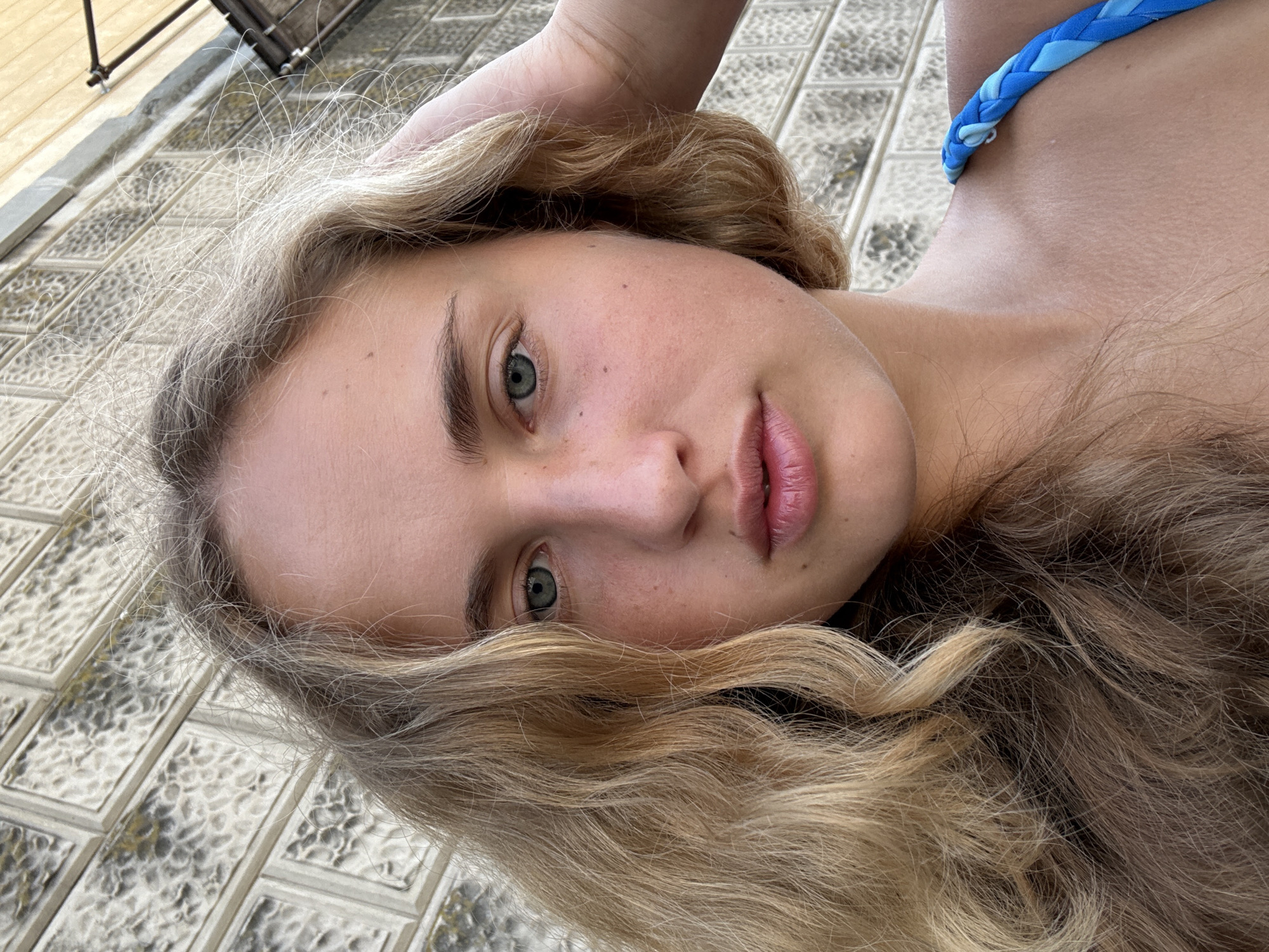 A close-up, selfie-style photograph captures a young woman with blonde, wavy hair and striking blue-green eyes against a textured, greyish-white brick wall. Her skin is fair with visible freckles. She is wearing a bright blue braided strap, likely from swimwear. Her left hand is raised, touching the side of her head, with her fingers splayed. The lighting suggests it is daytime. The brick wall has a repeating pattern of rectangular blocks with an irregular, almost pitted surface texture, common in older European architecture. The angle of the photograph is from a slightly low perspective, looking up towards the subject. The overall scene evokes a casual, outdoor setting.