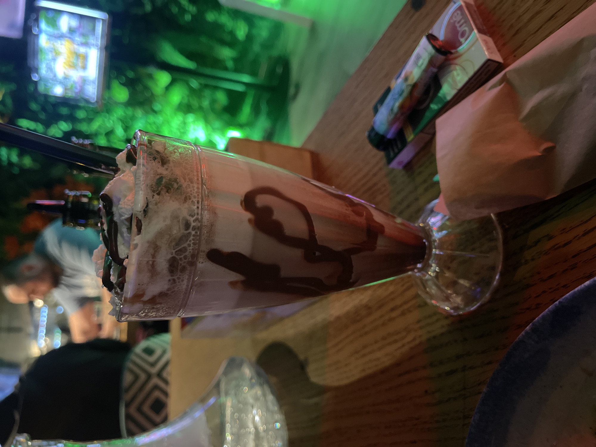 A medium shot of a sundae served in a tall, clear, glass-stemmed dish sits on a wooden tabletop, alongside a blue plate, a crumpled brown bag, and a colorful electronic cigarette with its packaging. The sundae is topped with whipped cream and drizzled with chocolate syrup. A dark straw is inserted into the sundae. The background is brightly lit with green light and features blurred figures of people and a screen displaying graphics. The image was likely taken indoors, possibly at night. While Sofia, Bulgaria cannot be definitively stated as the location from visual information, this location is presumed, based on image prompt.