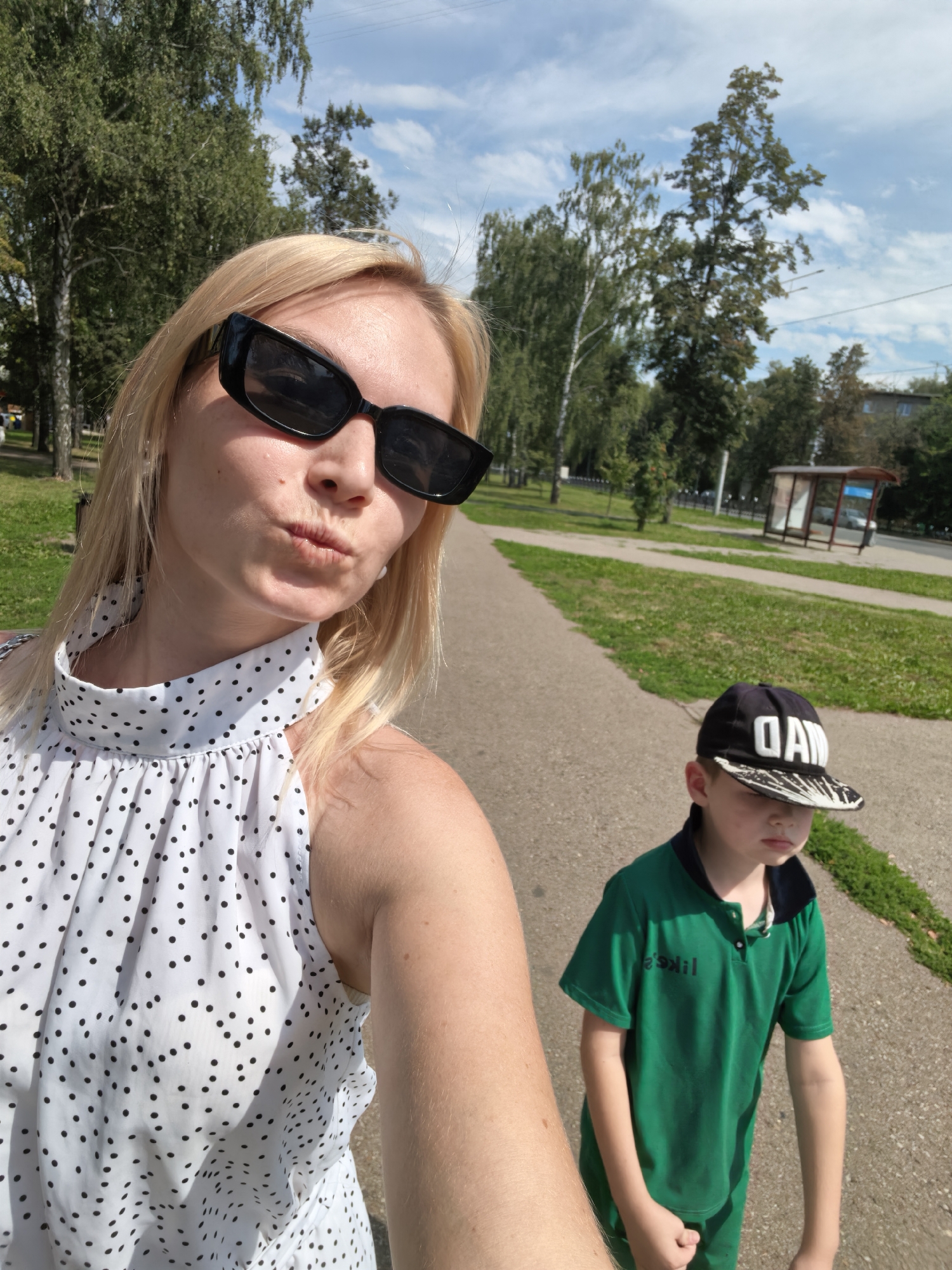 A blonde woman in sunglasses and a white polka dot top poses for a selfie, puckering her lips. Beside her, a young boy wearing a green polo shirt and a black baseball cap with white lettering walks away from the camera on a paved path. The background shows a park-like setting with trees, grass, and a bus stop. The scene is likely in Sterlitamak, Russia, given the context.