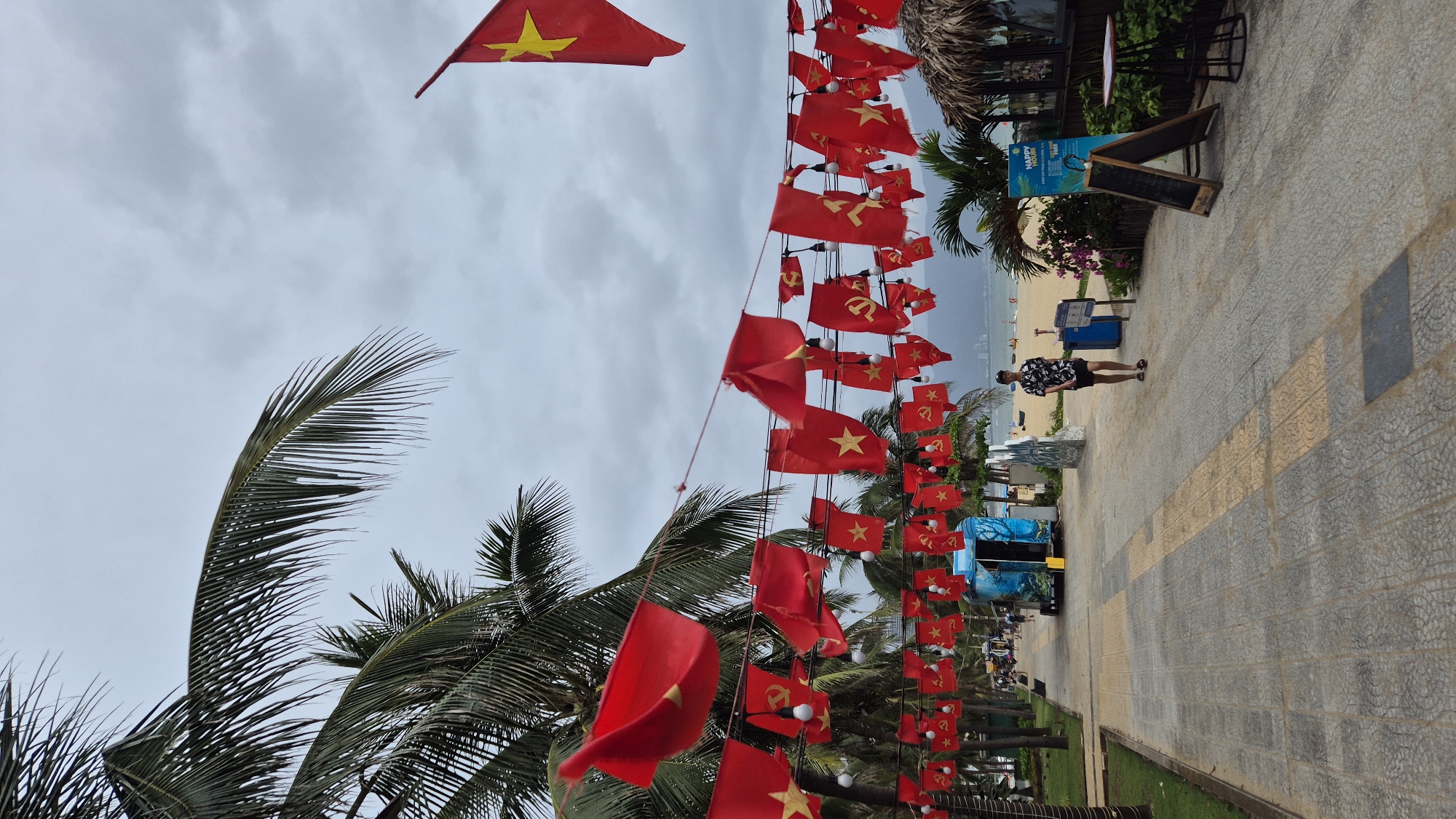 This image depicts a coastal scene in Da Nang, Vietnam, likely near My Khe Beach. A paved walkway runs parallel to the beach, lined with palm trees. Red flags with yellow stars, characteristic of Vietnam, are strung across the walkway at two levels, interspersed with other red flags featuring a hammer and sickle symbol, indicative of the Communist Party of Vietnam. A few bare lightbulbs are also visible on the lower string of flags.
In the foreground, a man wearing a patterned short-sleeved shirt and dark shorts walks along the path. To his right, a blue recycling bin and a wooden easel with a chalkboard are present. Further down the walkway, a blue mobile kiosk with an awning is visible.
The background reveals a sandy beach with a few scattered beachgoers and some boats. The ocean is visible beyond the beach, and a hazy mountainous coastline can be seen in the far distance under an overcast sky. The overall atmosphere suggests a public space decorated for a national or political event.
