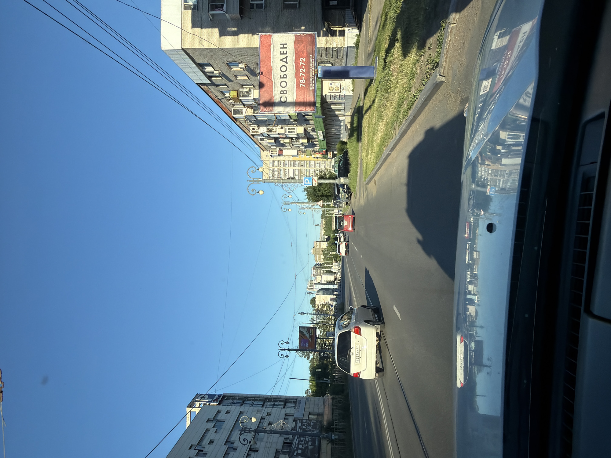 The image shows a view from a vehicle driving on a street in Khabarovsk, Russia. The street is lined with multi-story apartment buildings and some commercial establishments. On the right side of the road, a billboard displays the Russian word "СВОБОДЕН" (meaning "free") along with a phone number and a website address. Several cars are on the road, including a white hatchback in the foreground. The sky is clear and blue. The reflection on the car's hood shows buildings and other street elements. The overall scene depicts a typical urban streetscape in the city.