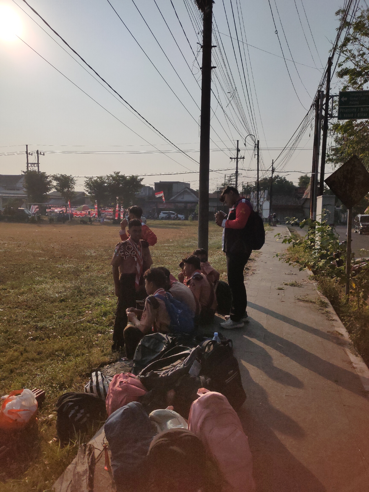 In the city of Ngasem, Indonesia, at dawn, a group of young people dressed in scout uniforms are gathered on the side of a paved path. Some are sitting on the grass, while others stand. They appear to be resting, perhaps before or after an event. Luggage is piled up in the foreground. In the background, there are buildings, trees, and a field with red and white banners. Power lines crisscross the sky, indicating the presence of urban infrastructure. The sun is low in the sky, casting long shadows.