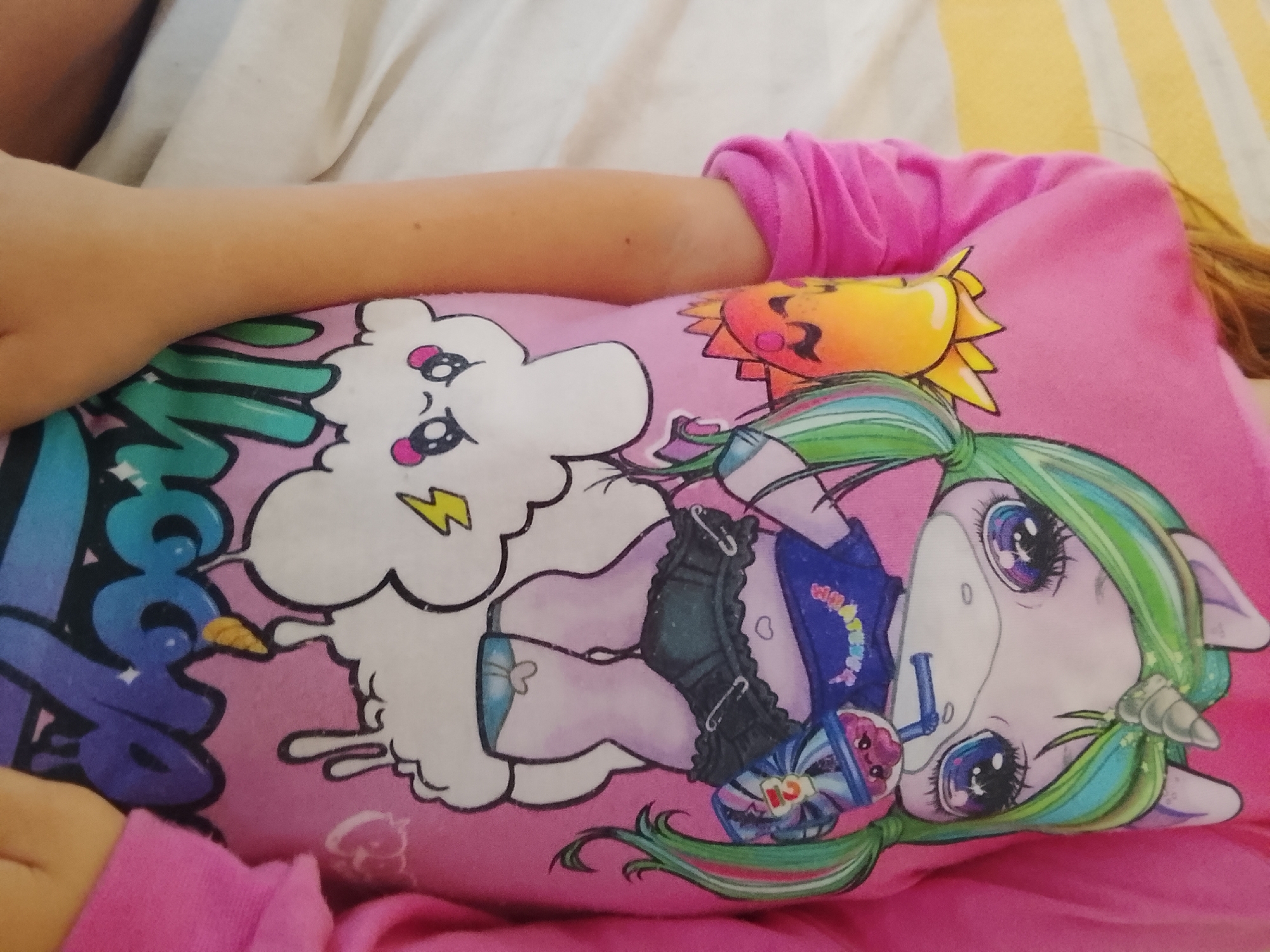 A child is wearing a pink shirt with cartoon graphics on it, including a white cloud with a lightning bolt, a cartoon sun, and a unicorn with green and teal hair, holding a drink. The child is lying in a bed with white and striped yellow sheets. The location is not discernible in the image.