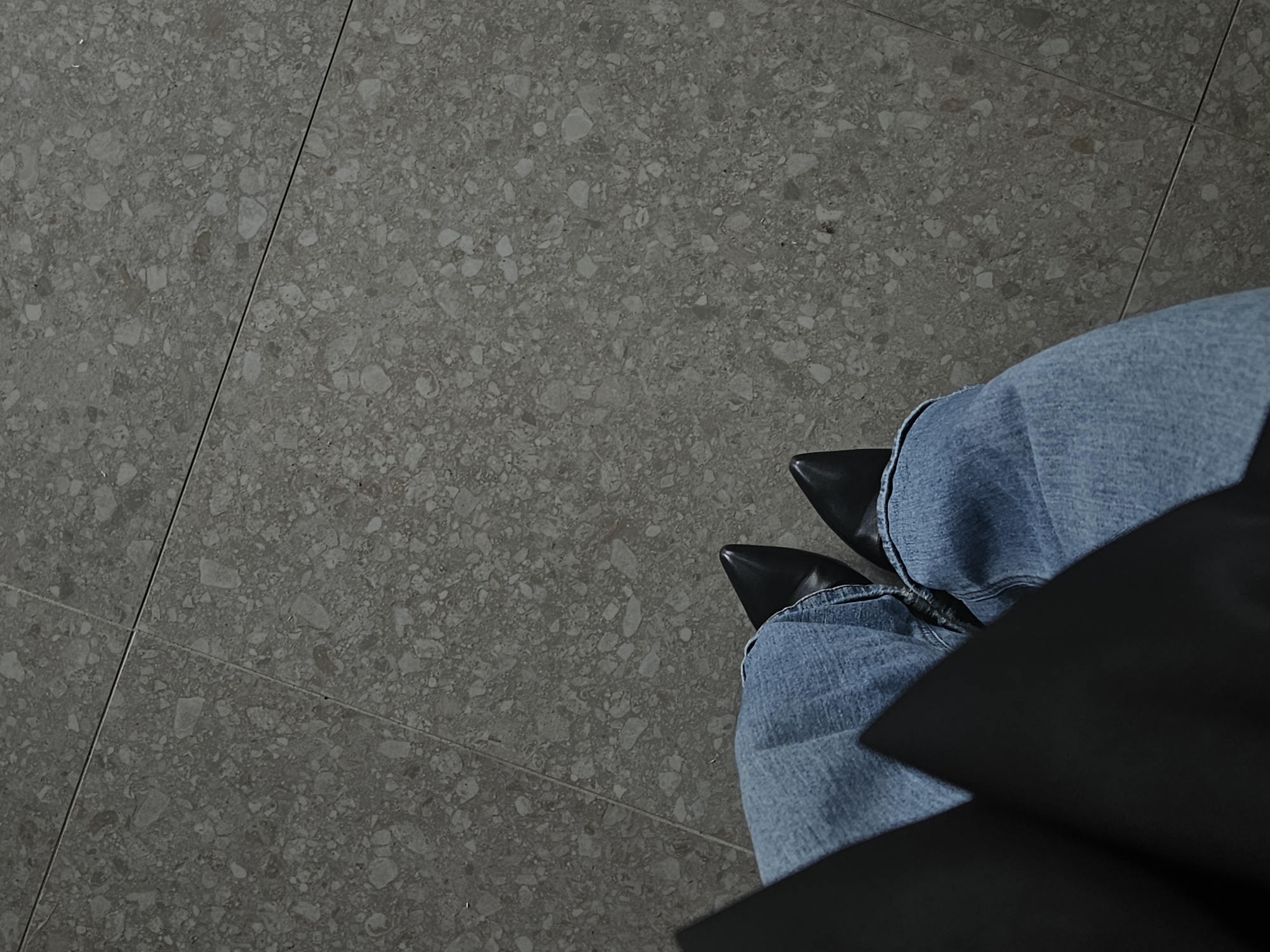 The image, captured from a high-angle perspective in Chaikovsky, Russia, features a person's lower body against a tiled floor. The main subjects are a pair of legs dressed in blue denim jeans, which are slightly bunched around the ankles. At the bottom of the jeans, black, pointed-toe shoes or boots are visible. A dark, solid object or surface, possibly part of a chair, bag, or other item, partially obscures the right side of the person's lower legs and shoes, extending from the bottom right corner.
The setting is an indoor space, characterized by the flooring, which consists of large, rectangular grey tiles. These tiles have a distinct terrazzo or aggregate finish, featuring a mottled pattern of various light grey, white, and subtle beige chips embedded within a darker grey matrix. Dark grout lines demarcate the individual tiles, creating a grid pattern.
No specific activity is depicted beyond the person's static posture, suggesting they are either standing or sitting and looking down. The time of day, weather, and emotional state are not discernible from the image. There is no visible text.